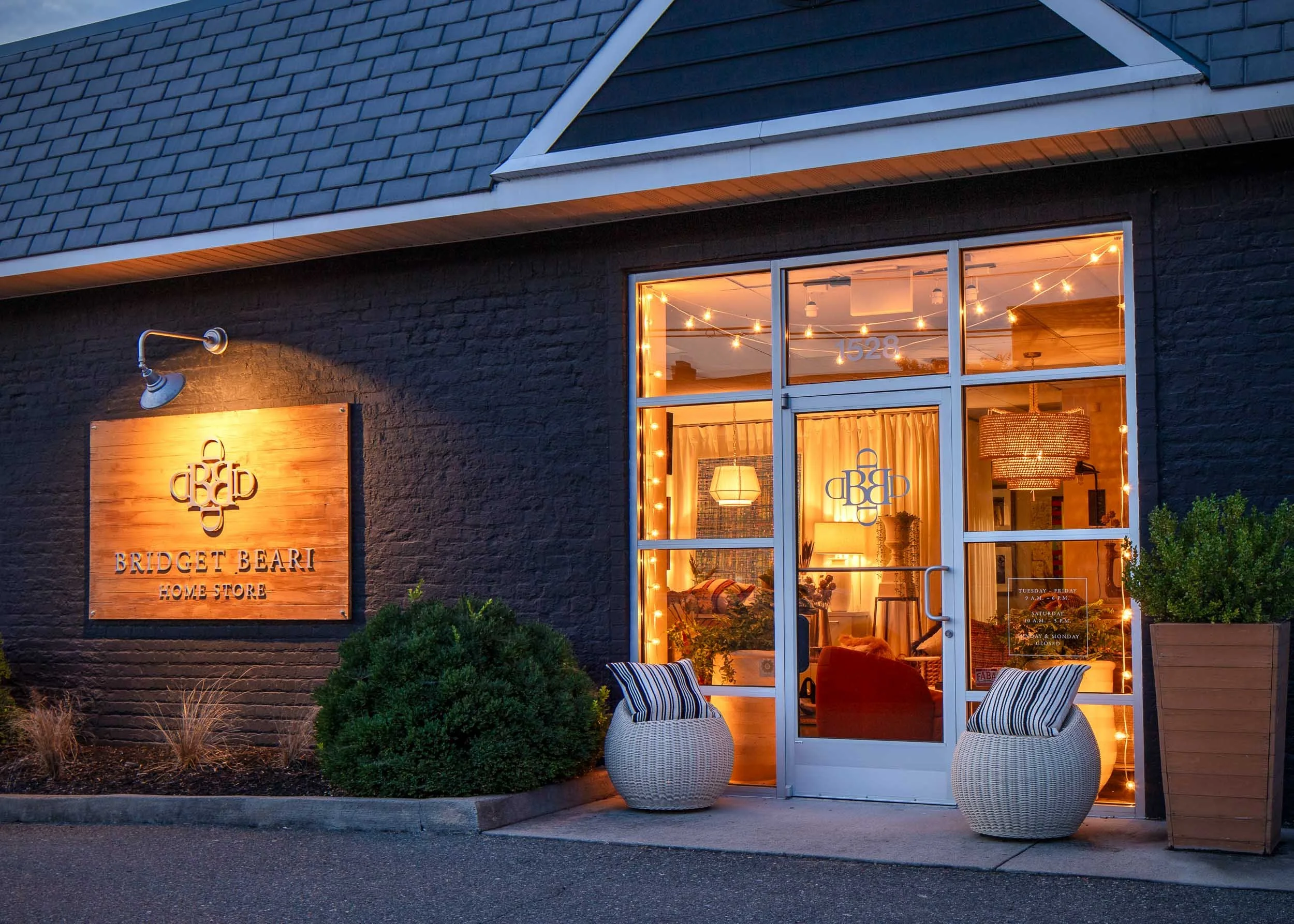 Exterior view of Bridget Bearl Home Store illuminated at dusk, showing a black brick wall, a wooden sign, a glass door with lights inside, two wicker chairs with striped cushions, potted plants, and a large window.