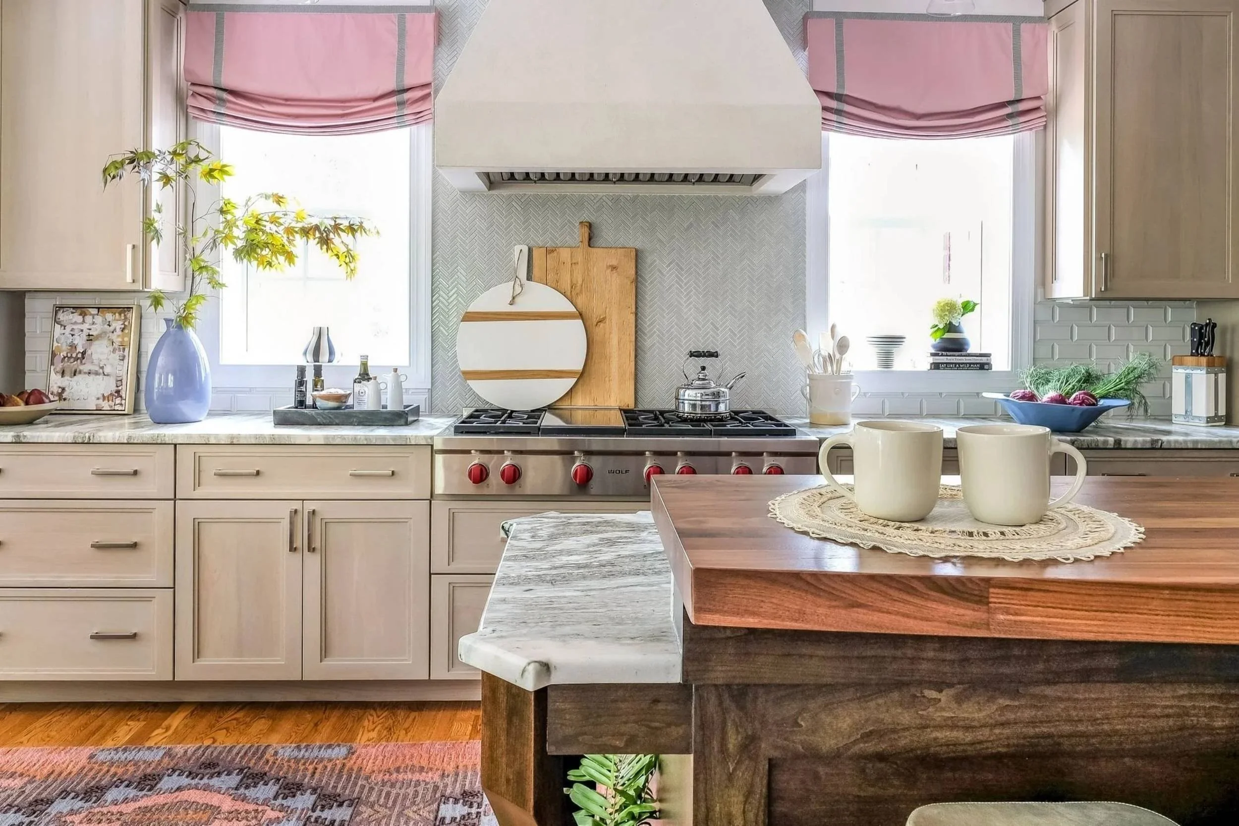 An island sits in the foreground of a kitchen stove, with light wood cabinets and pink draperies on the two windows on either side of the overhead vent. Two cream mugs rest on a doily on the island.