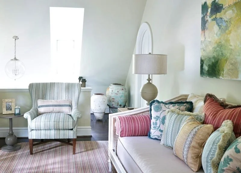 A cozy living room with a striped armchair, a sofa with colorful throw pillows, a floor lamp, a side table with decorative jars, and an abstract painting on the wall.