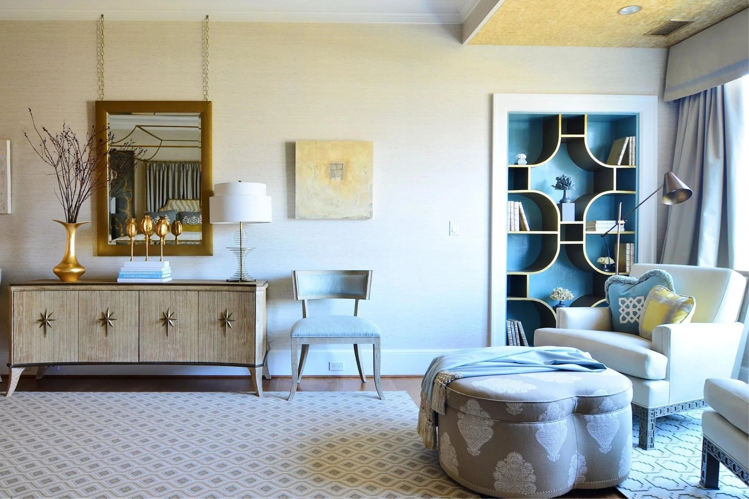 In a calm, yellow-ish cream room, a large gold mirror overtop a chest sits to the left of a built in book case with blue panelling. A cozy oversized chair and ottoman sit in the right corner, next to the window.