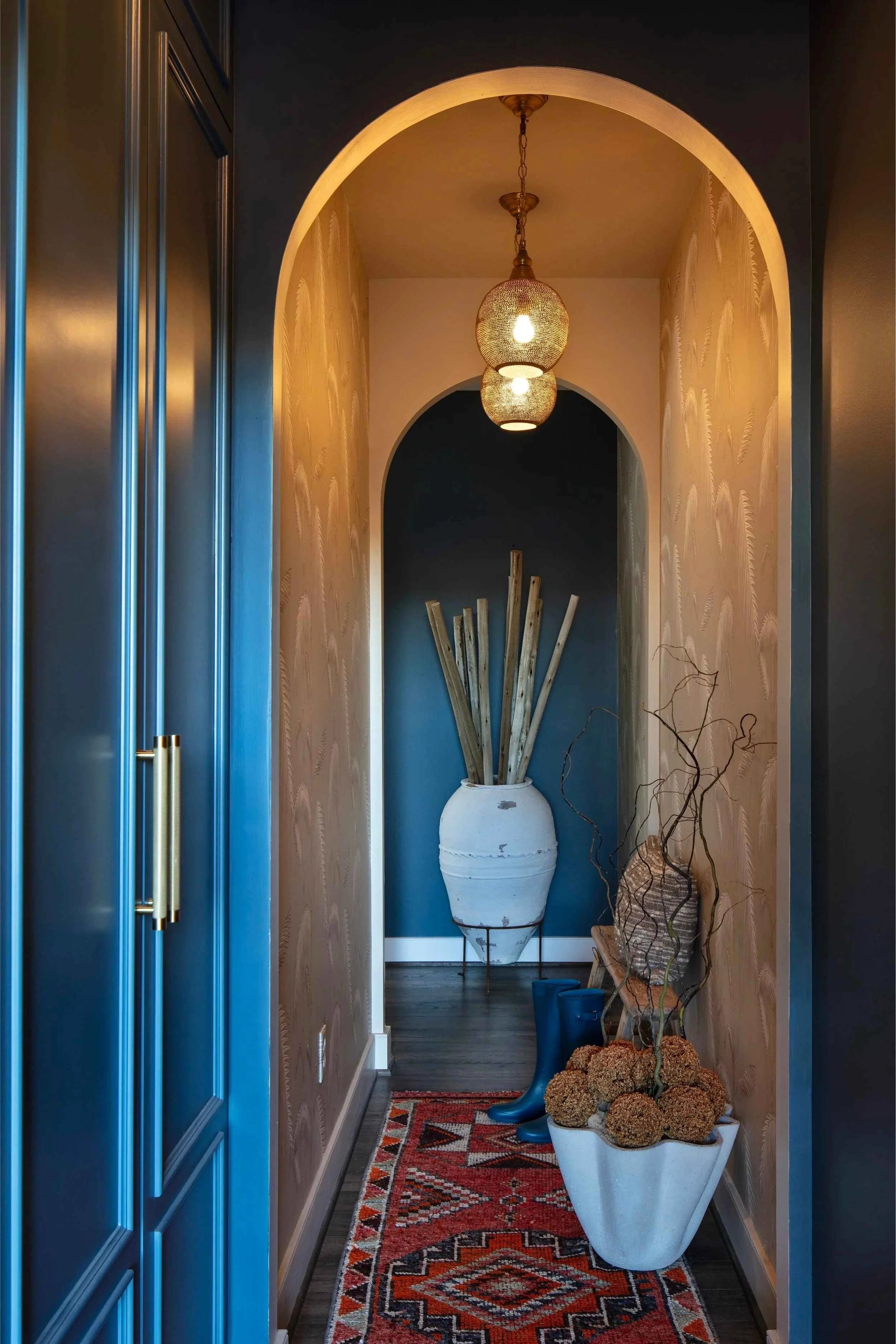 A rich dark blue leads into a beige, wallpapered hallway in this entry room. Two round light fixtures hang, and a large vase with drift wood sits at the back of the hallway.