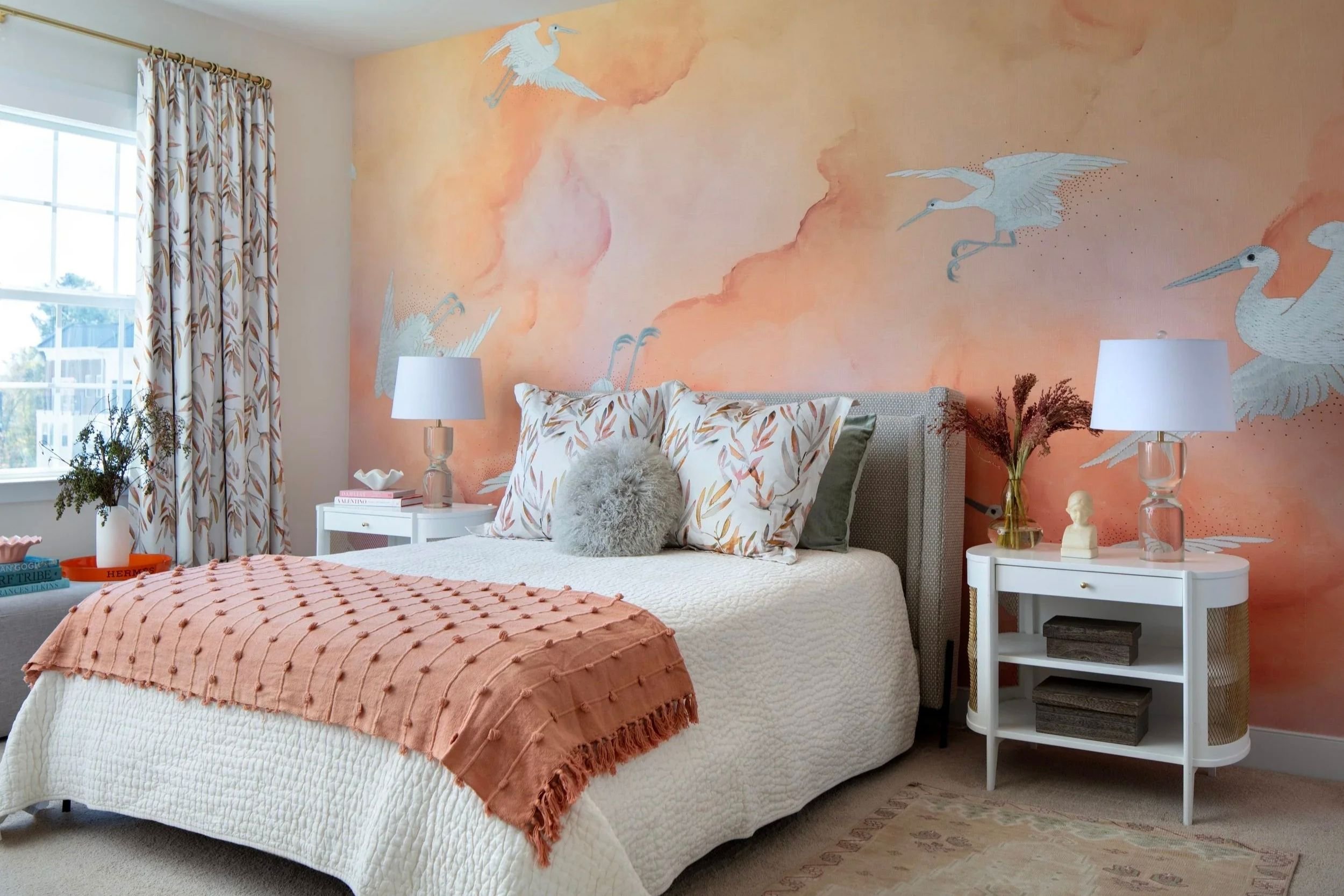 A queen-sized bed is shown against a coral-orange wall with white cranes on it. The bed contains coral pillows and a throw, and the two white bedside tables have various accessories along with two glass lamps. A window is shown on the left.