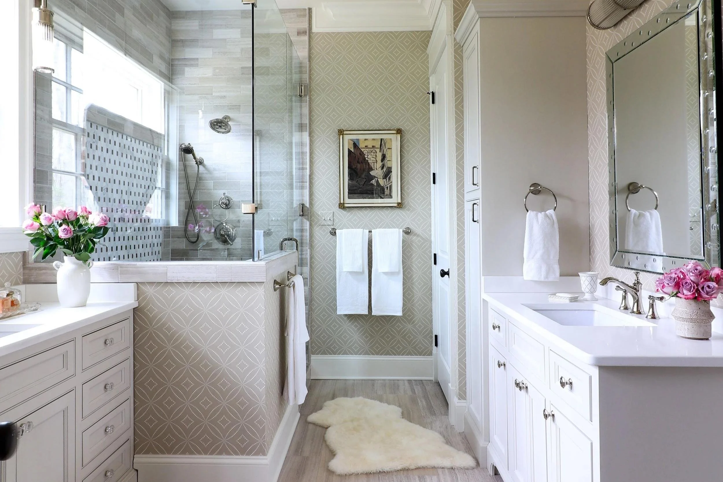 In the background of a main bathroom, a shower with a glass encasing is shown on the left, while cabinets are shown on the right. In the foreground, two sinks are across from each other on the left and right side. Walls are wallpapered beige pattern.