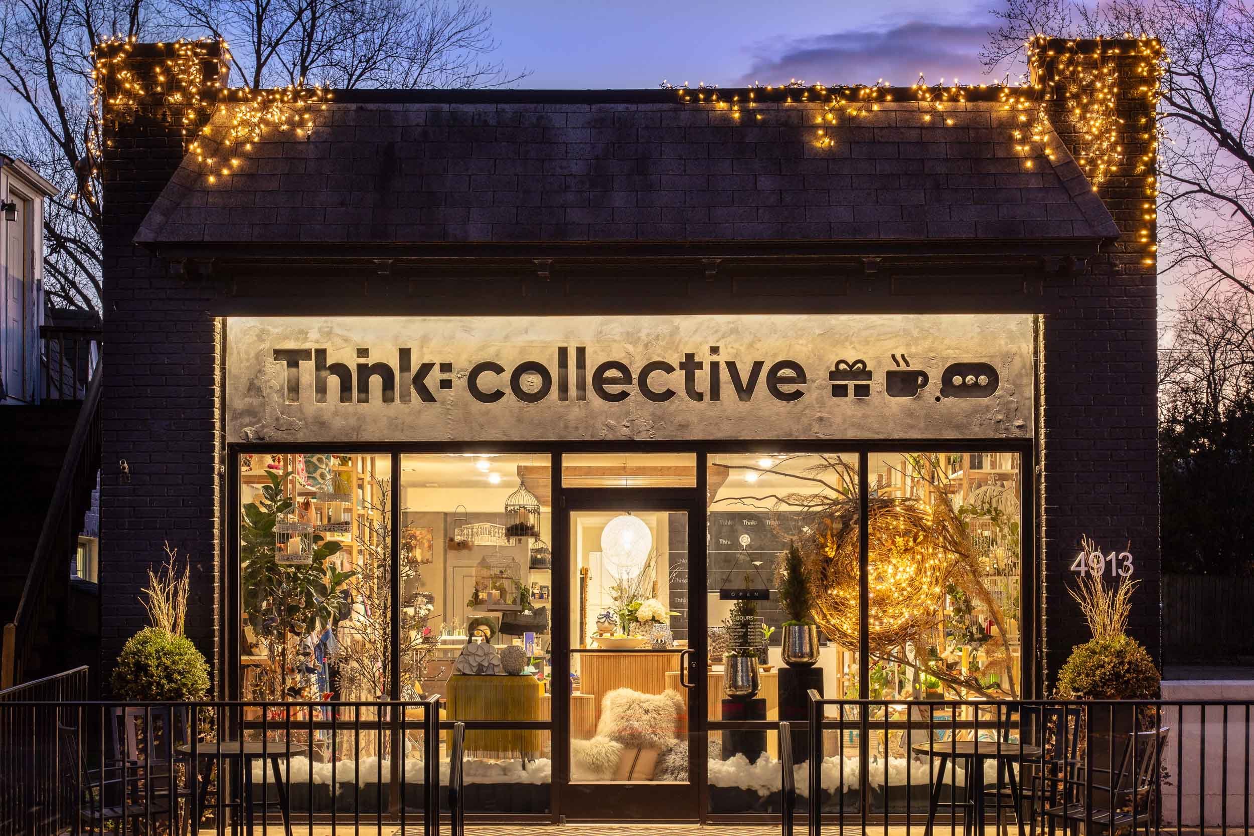The storefront of a shop called 'Think:collective' at dusk, decorated with Christmas lights along the roofline, with large windows displaying interior decor including plants, ornaments, and comfortable furniture.