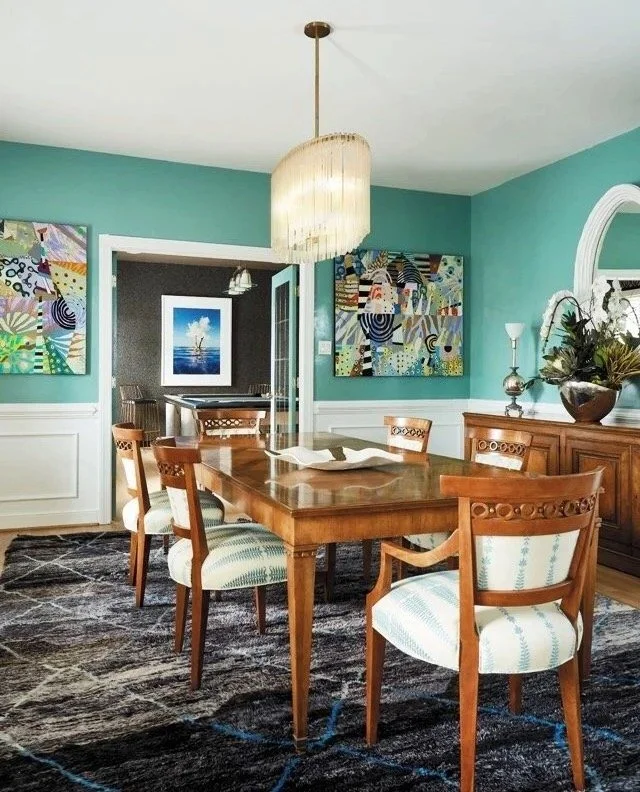 A dining room with a wooden table and 6 upholstered chairs sits beneath a fringed chandelier. The walls are painted a bright mint blue, with abstract art and a mirror on the right most wall. There is a dark, black and blue area rug beneath the table.