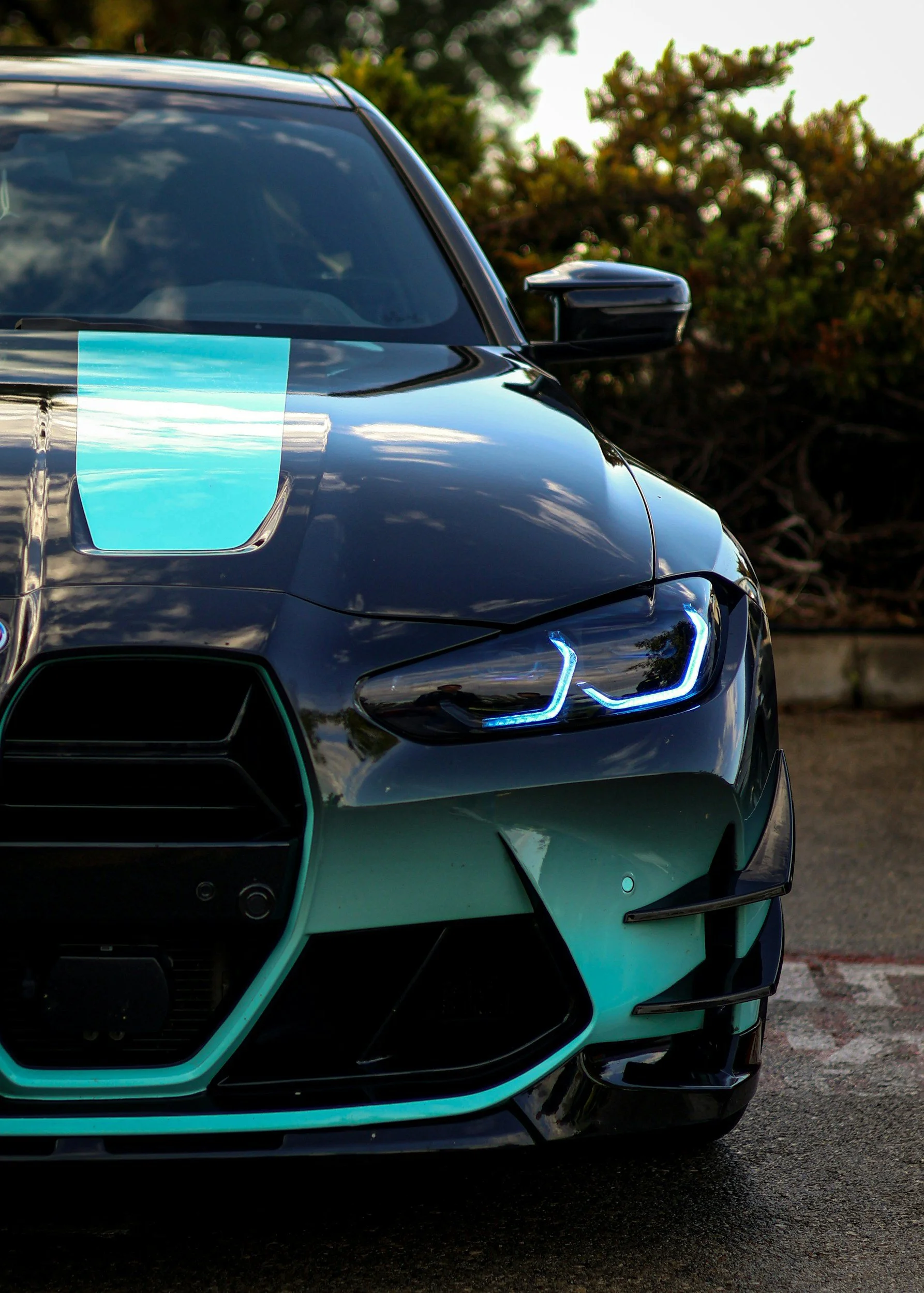 Close-up of the front of a sleek, black sports car with illuminated blue headlights and a teal accent on the front grille, parked outdoors with trees in the background.