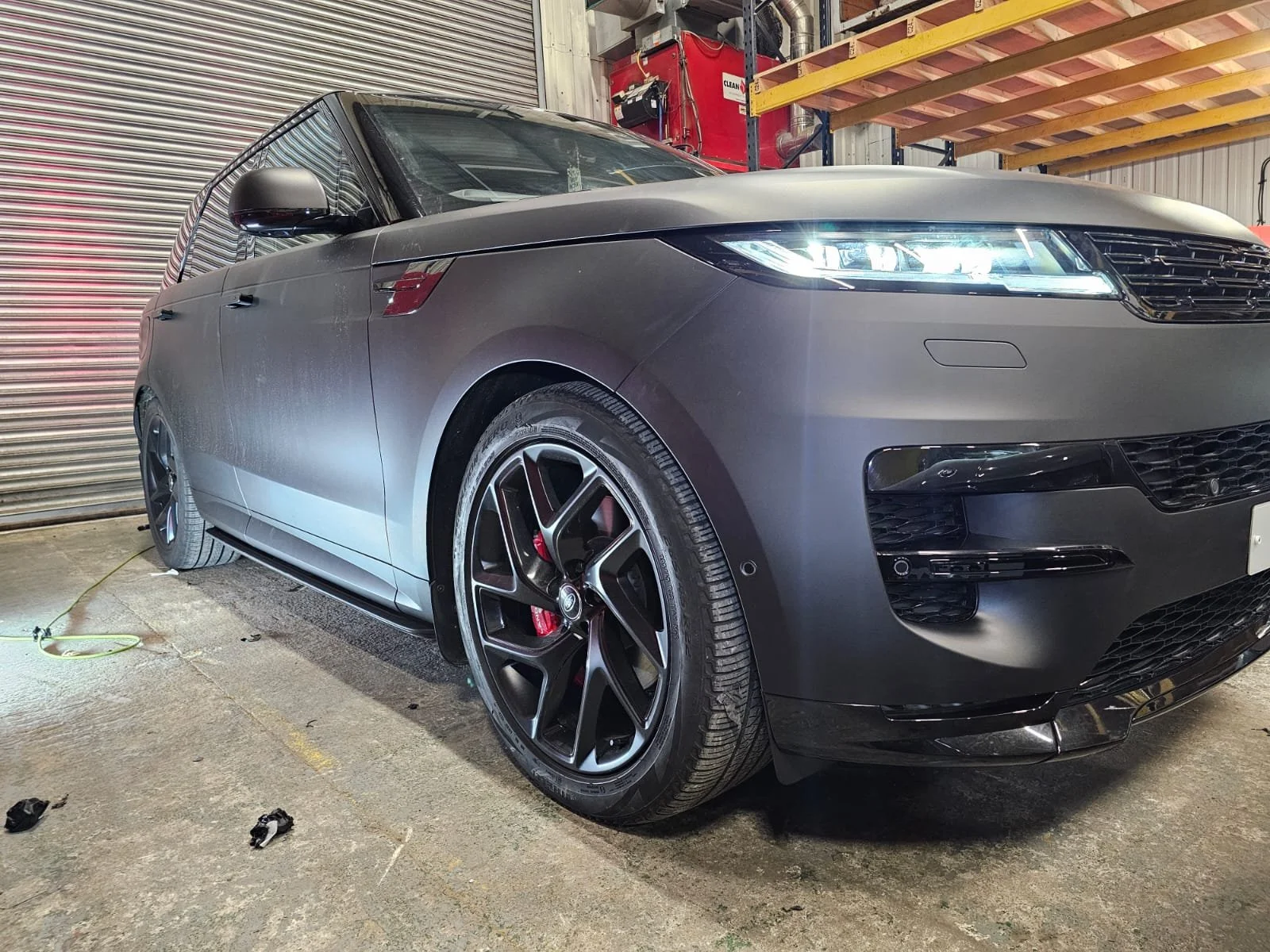 A black sedan car parked inside a garage with a concrete floor, partially visible roll-up door, yellow beam, and red equipment in the background.