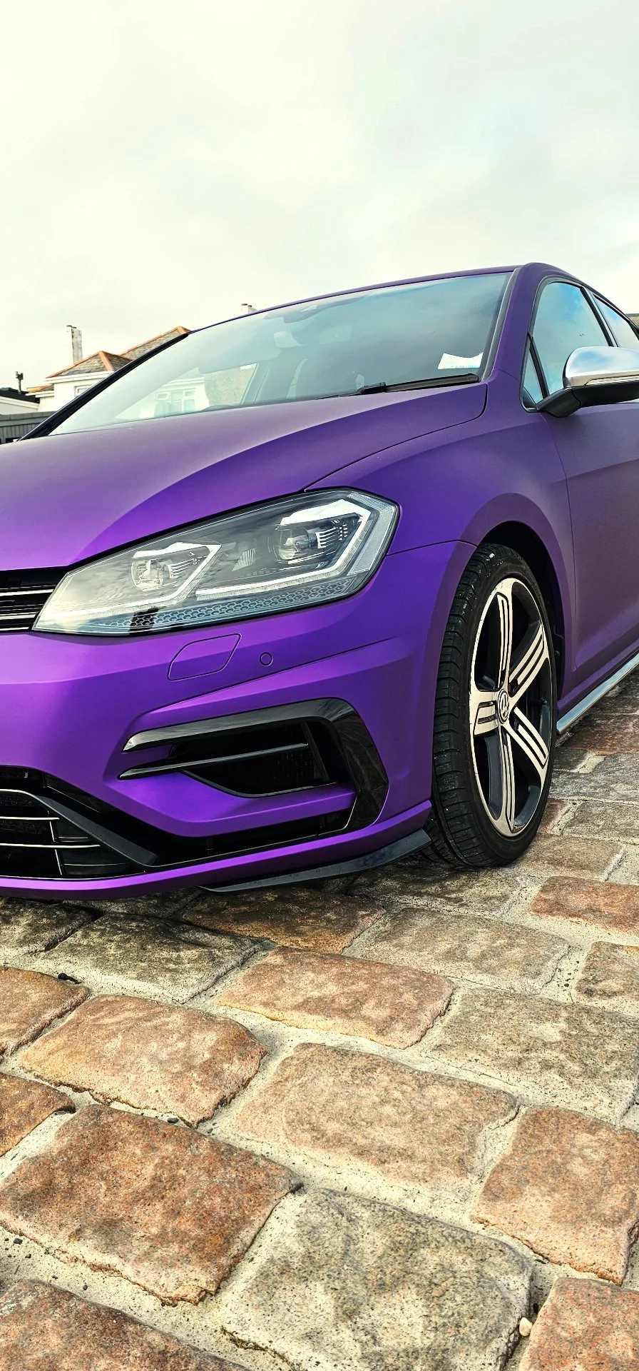 A purple modern car parked on a cobblestone driveway. Only the front portion of the car is visible, featuring sleek headlights and stylish alloy wheels.
