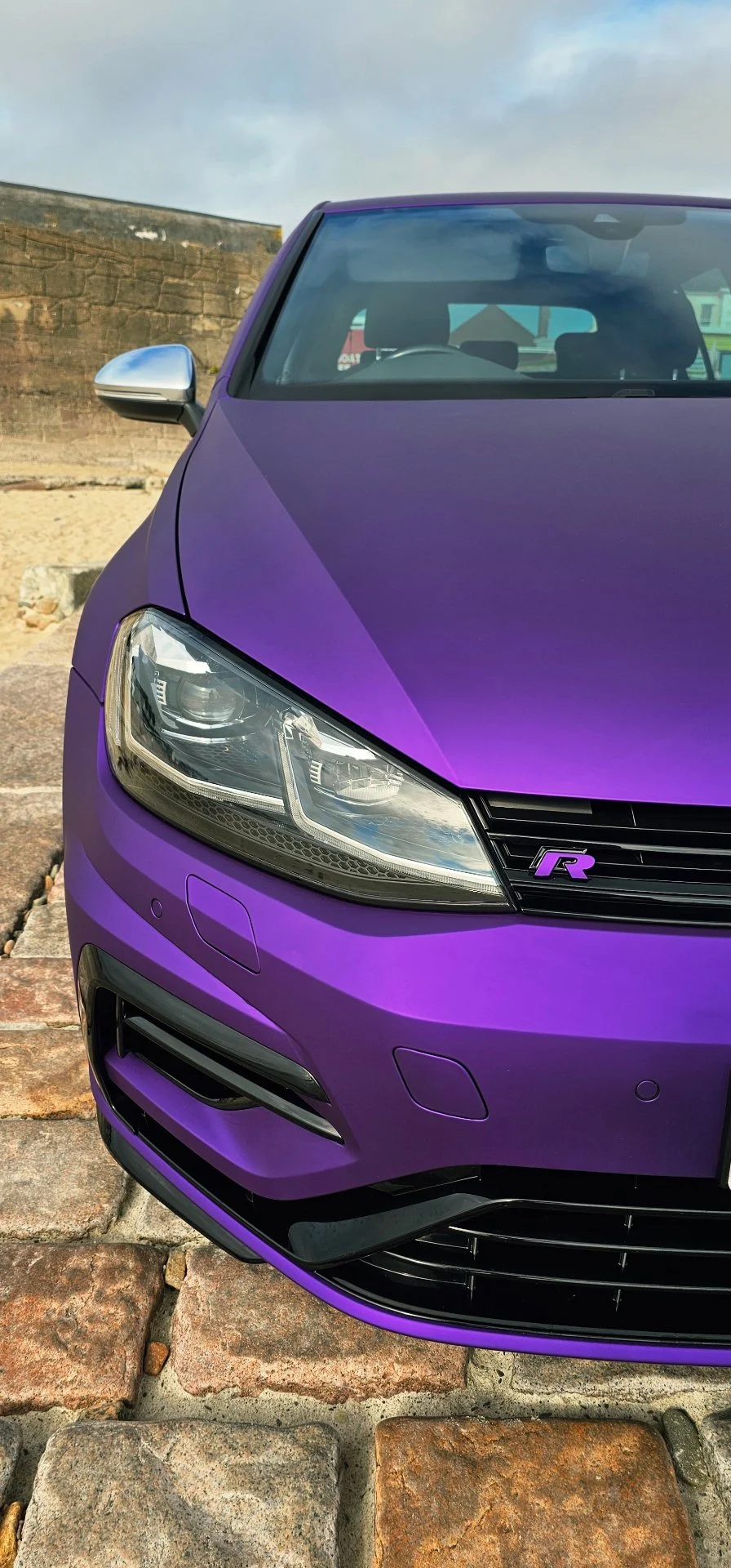Close-up of a purple sports car with a sleek front design, modern headlights, black grille, and an 'FR' badge on the front grille, parked on cobblestone pavement outdoors.