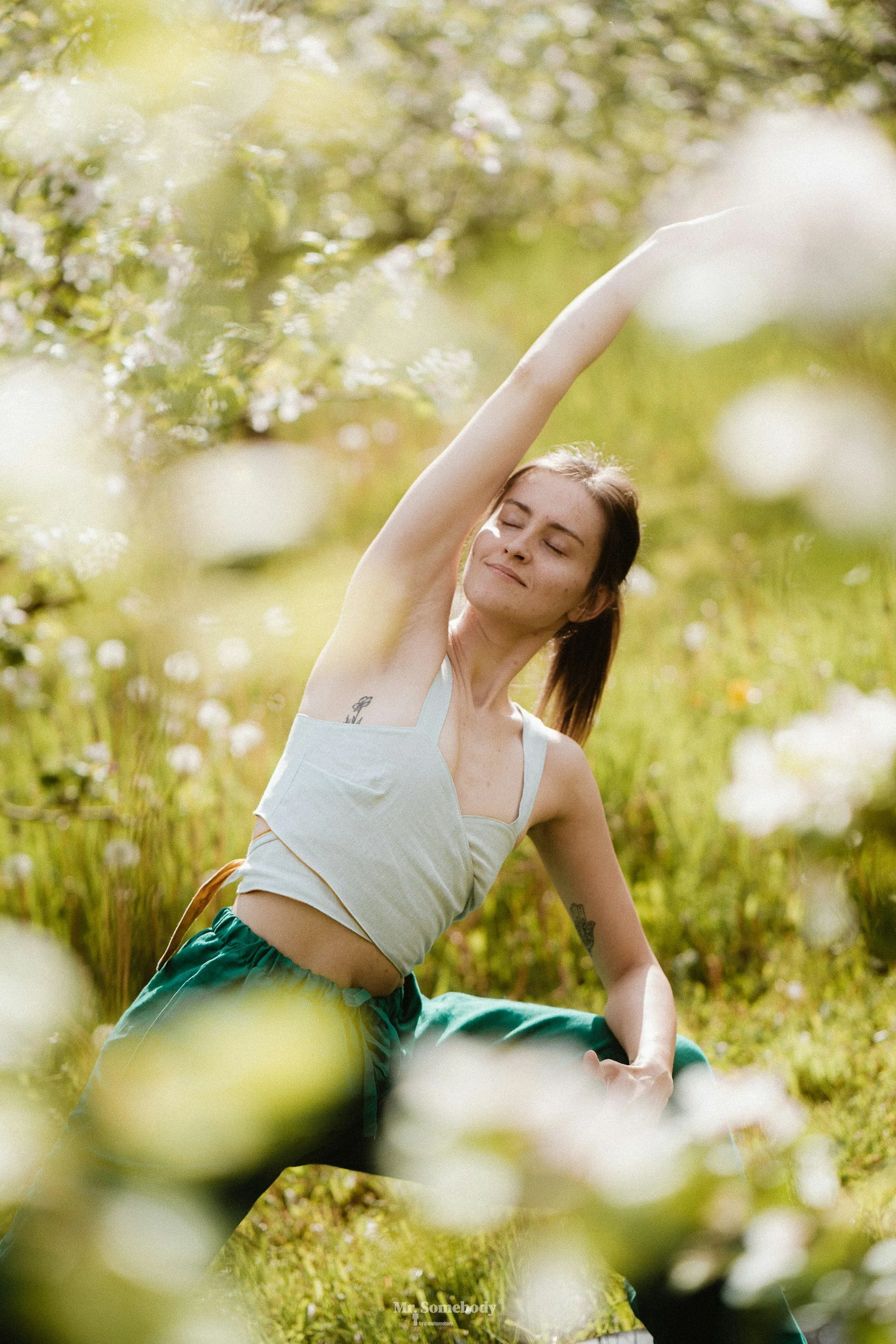 A young woman with closed eyes and a peaceful expression practicing yoga outdoors in a sunny, green, flower-filled field, raising one arm overhead.