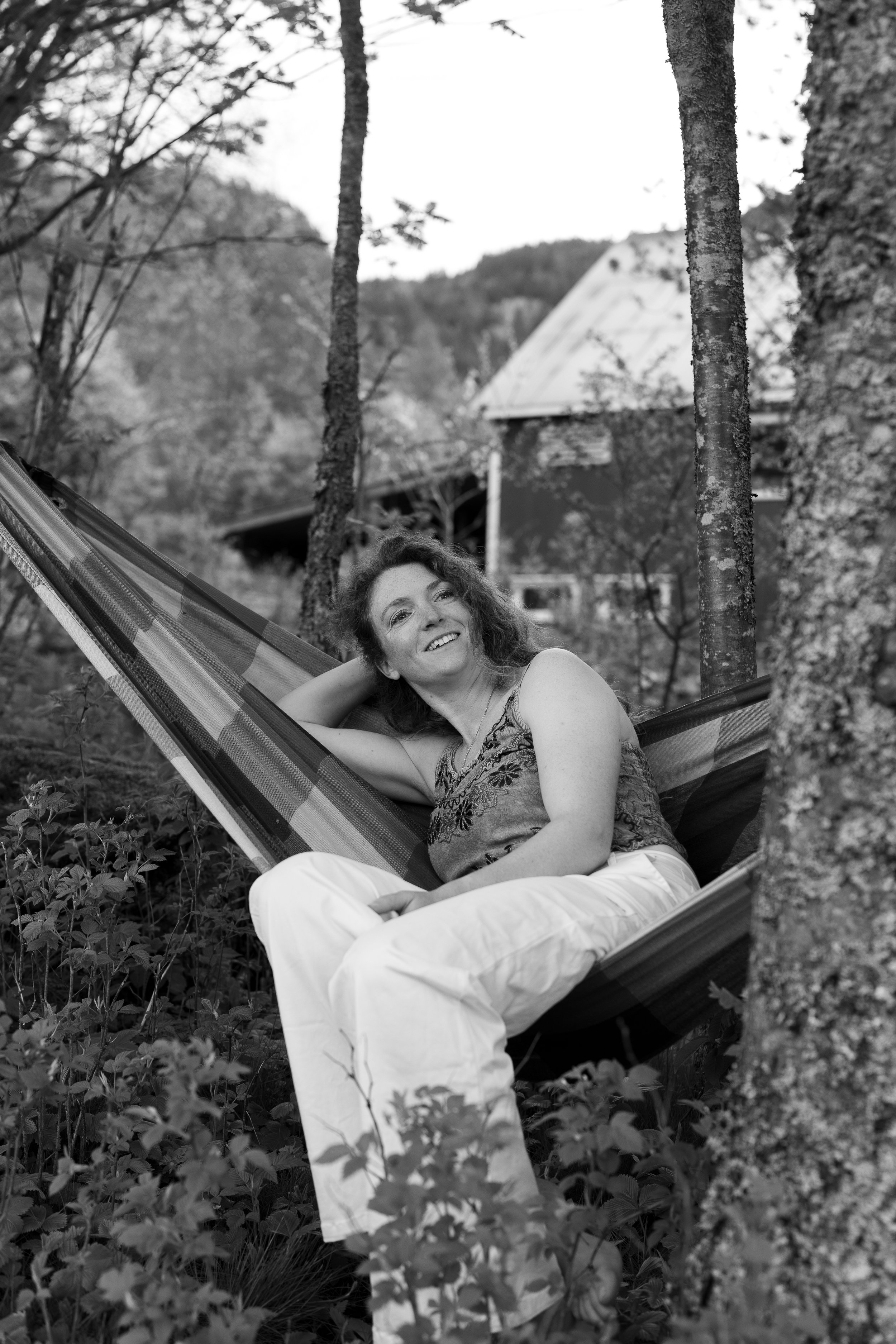 A woman lies comfortably on a hammock in a wooded outdoor setting, smiling at the camera with trees and a house or cabin in the background.