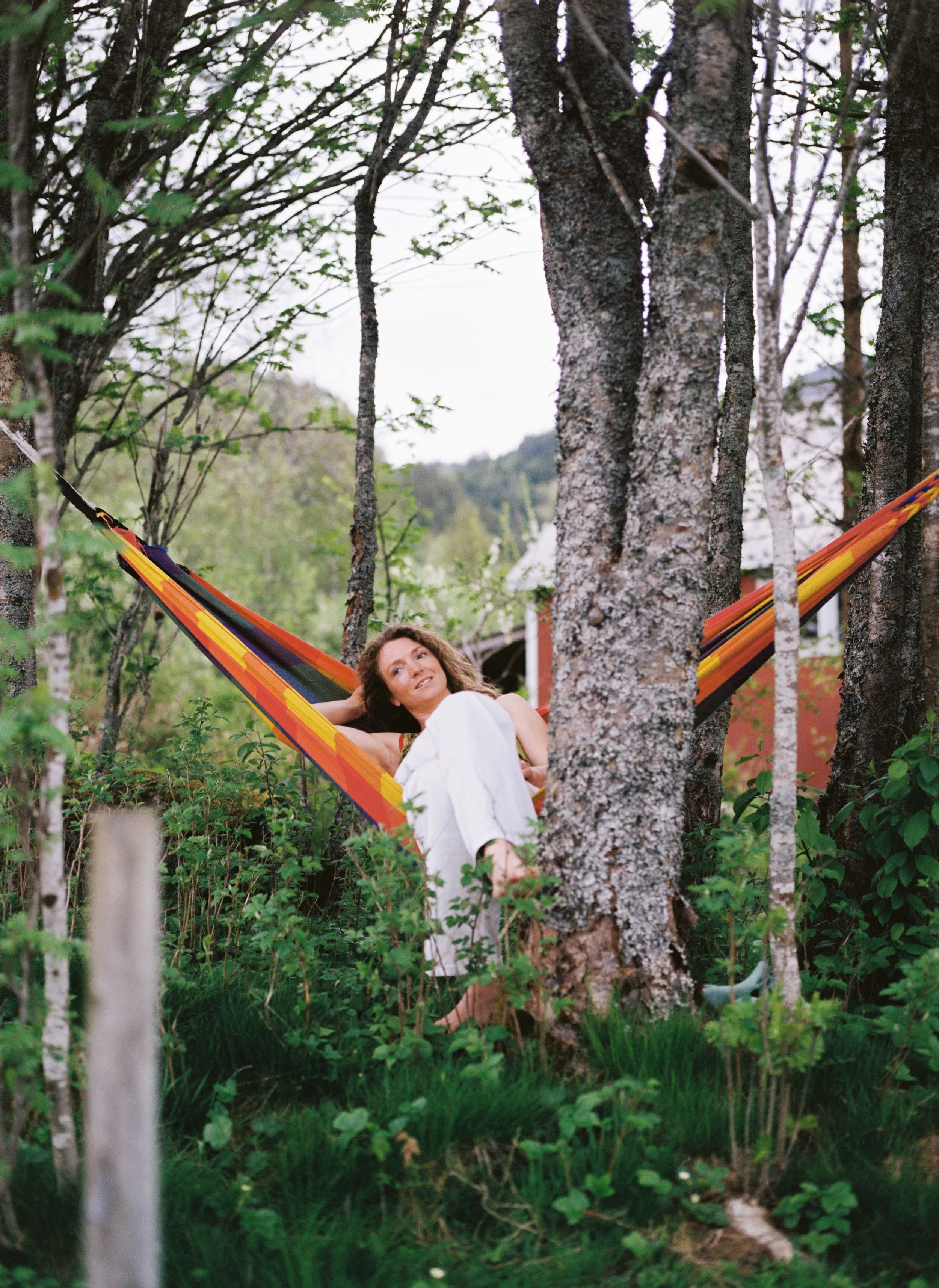A woman with curly brown hair relaxing in a colorful striped hammock tied between two trees in a lush green wooded area.