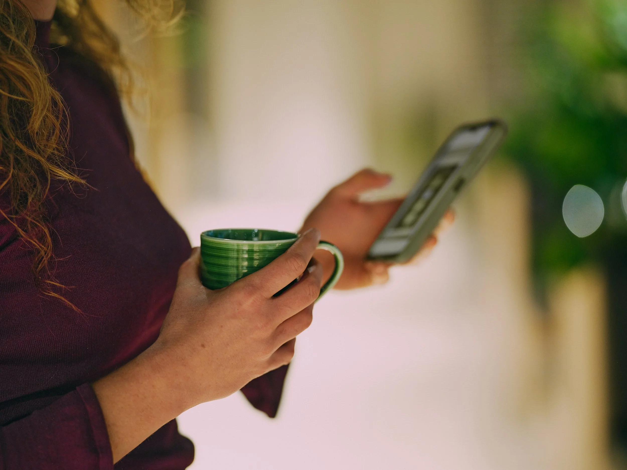 A woman with wavy hair holding a smartphone in one hand and a green and white striped mug in the other, standing indoors with a blurred background of green plants.
