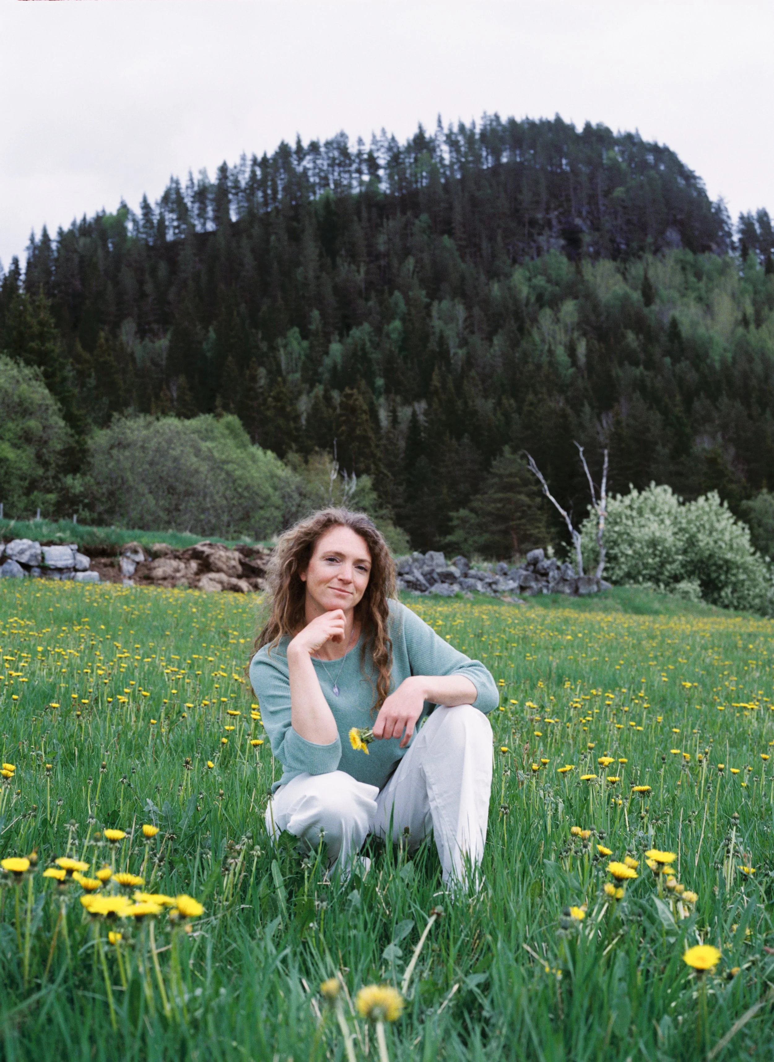 A woman with curly hair sitting in a field of yellow dandelions, with a wooded mountain in the background.