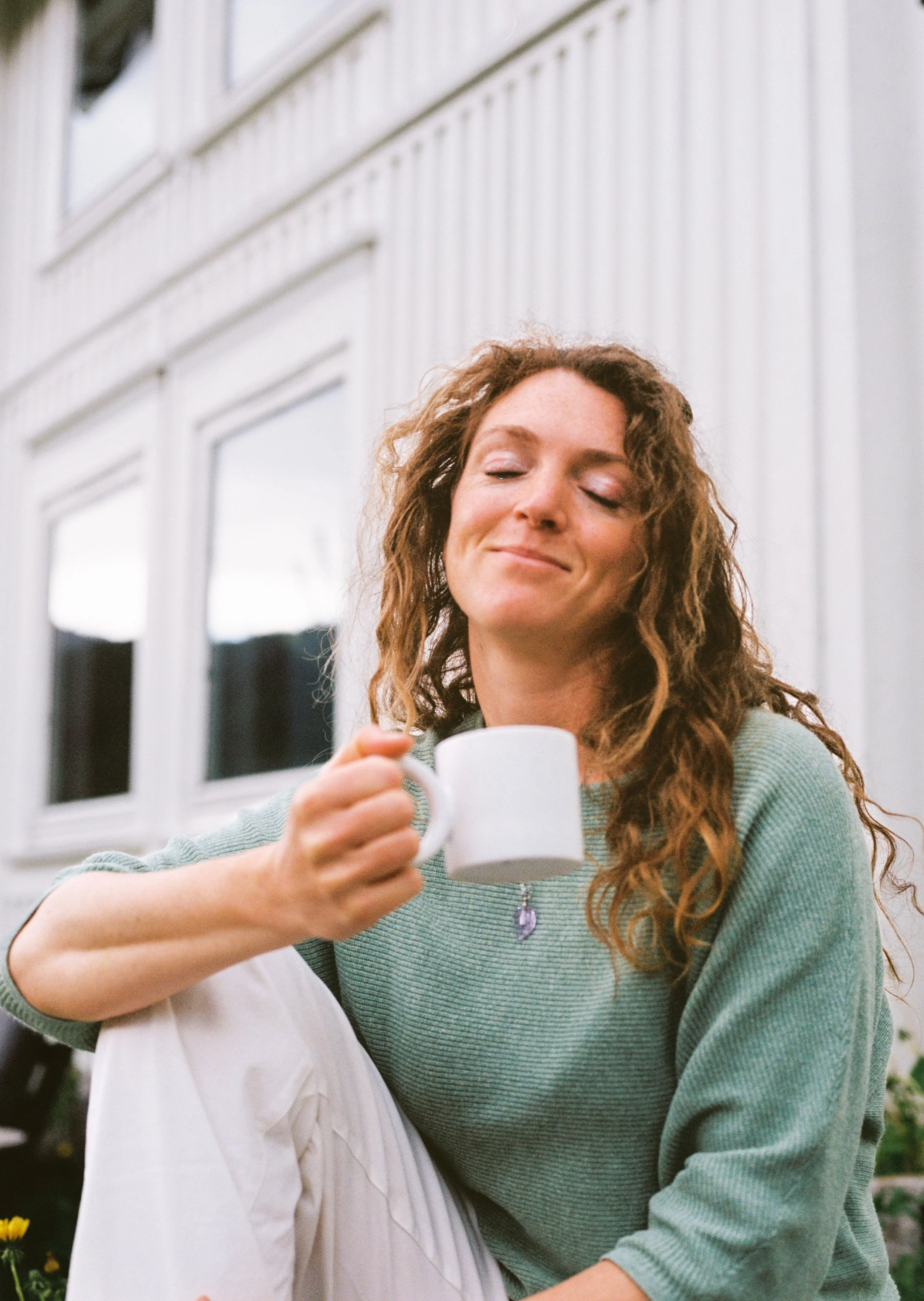 A woman with curly hair and a light blue sweater smiling with her eyes closed, holding a white mug, standing close to a person in a white shirt outdoors with a building and windows in the background.