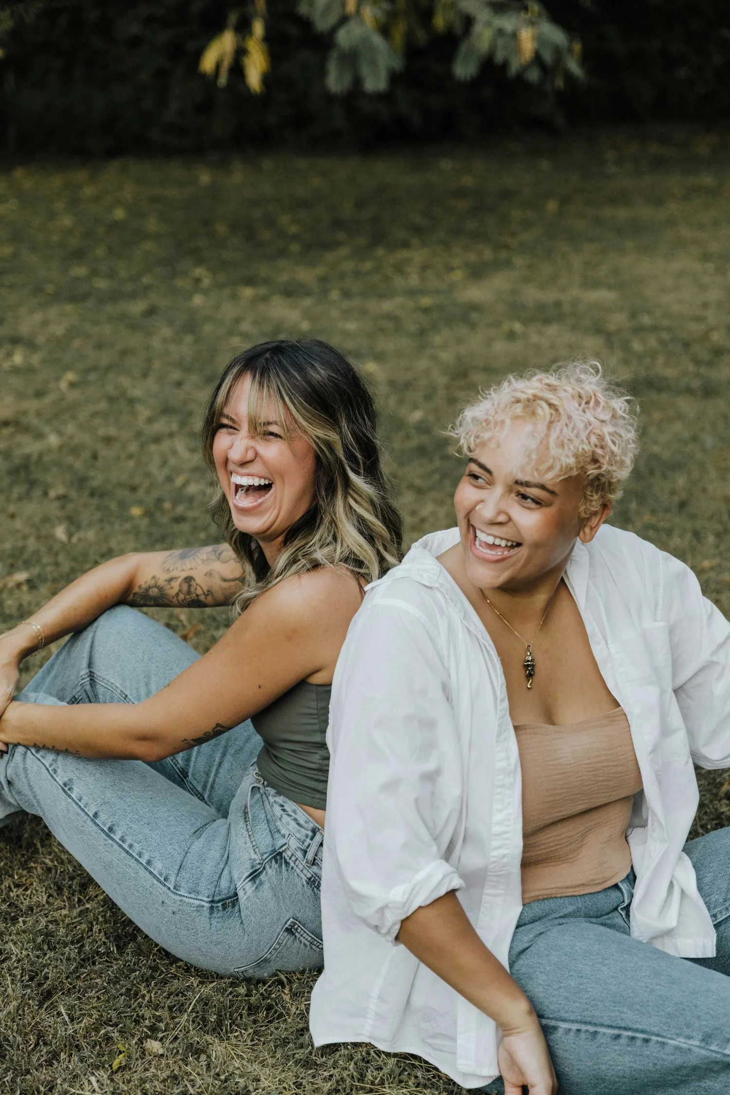 Two women sitting on the grass, laughing and smiling outdoors.