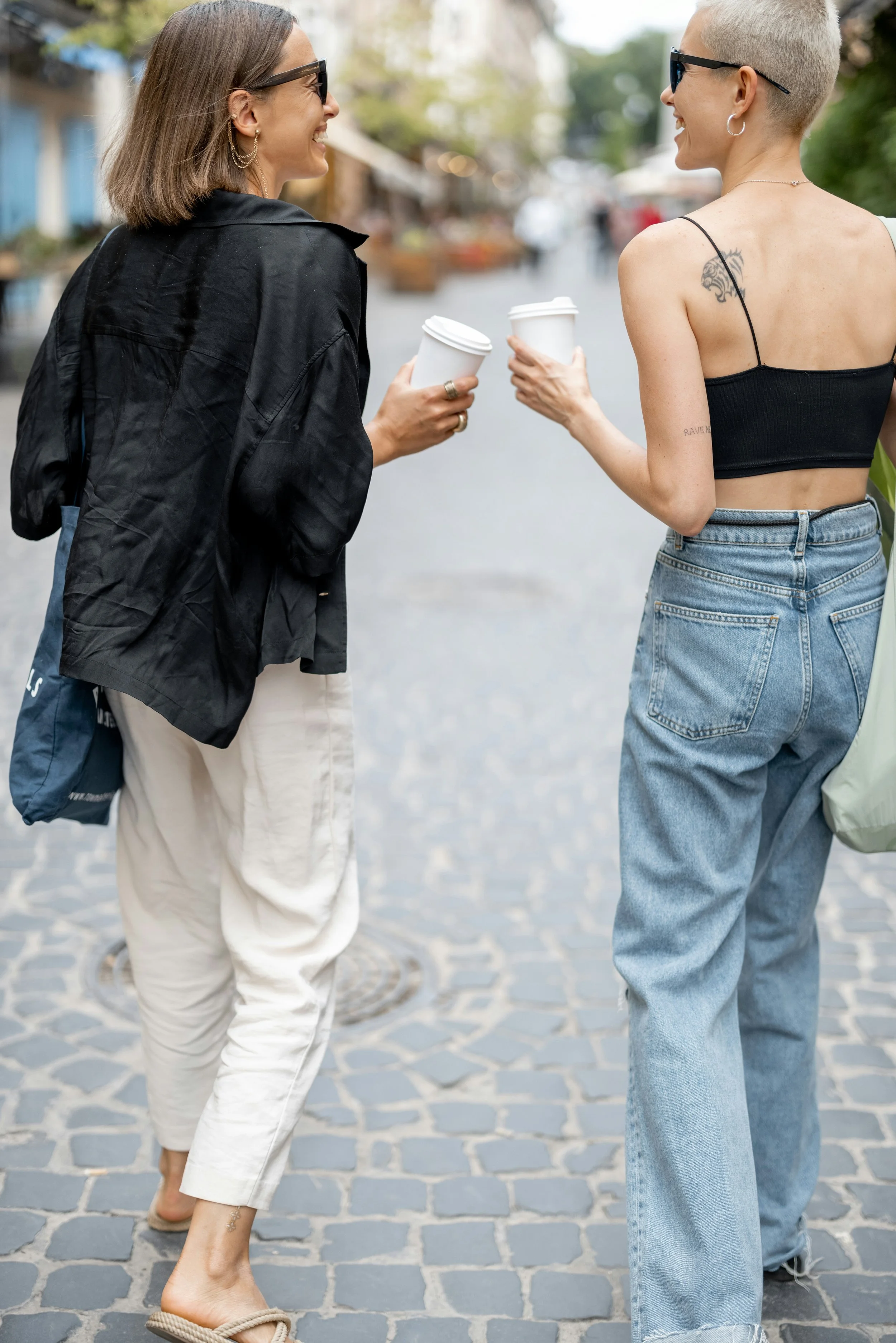 Two women walking and talking on a cobblestone street, each holding a to-go coffee cup, smiling and enjoying conversation.