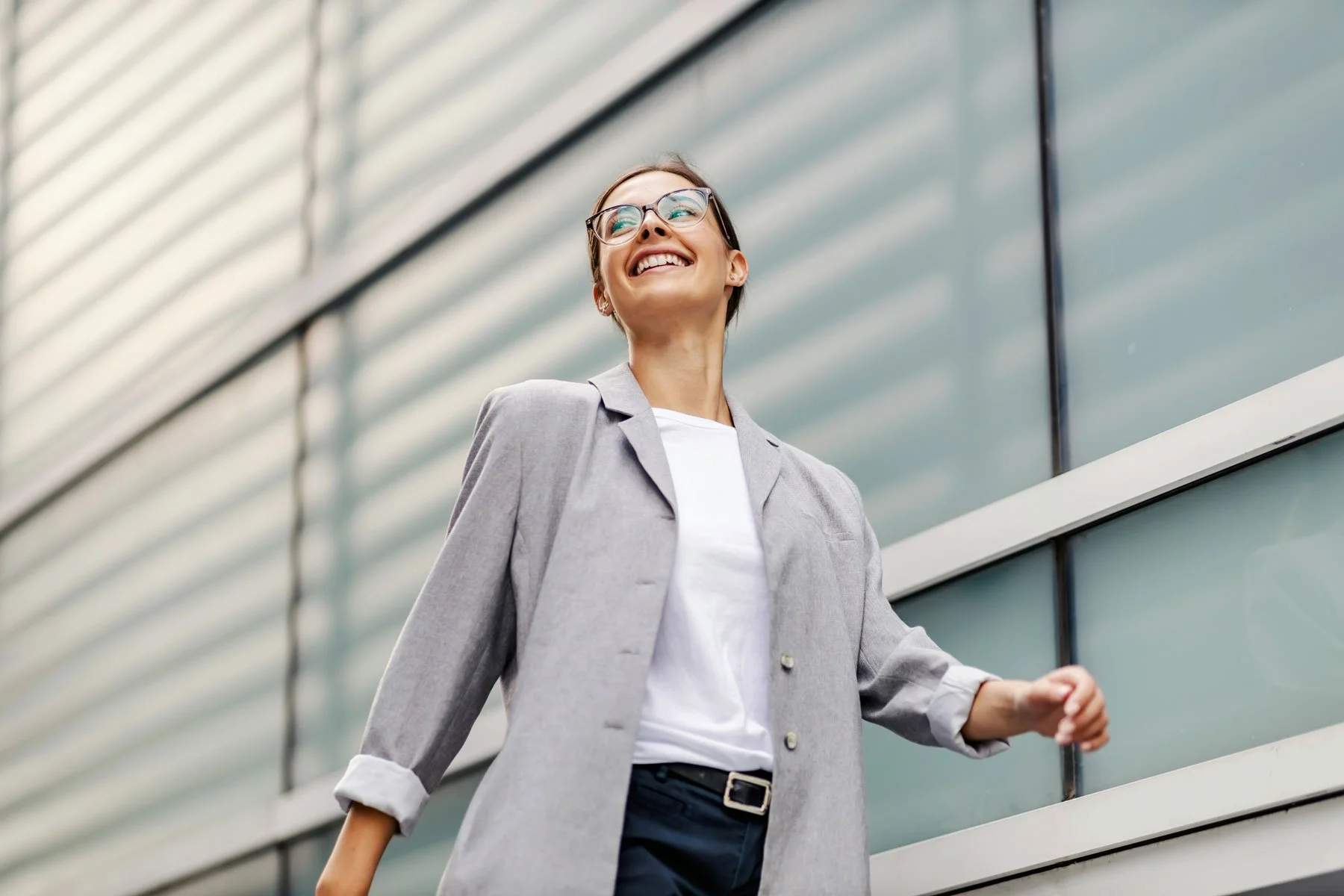 Smiling woman in business attire with glasses walking outside modern glass building.