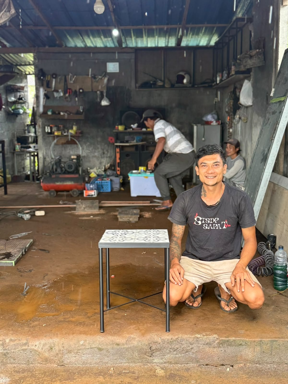 En man med tatueringar på armarna sitter på knä framför ett litet bord i ett slitet garage, medan andra personer arbetar i bakgrunden. Mannen ler mot kameran.