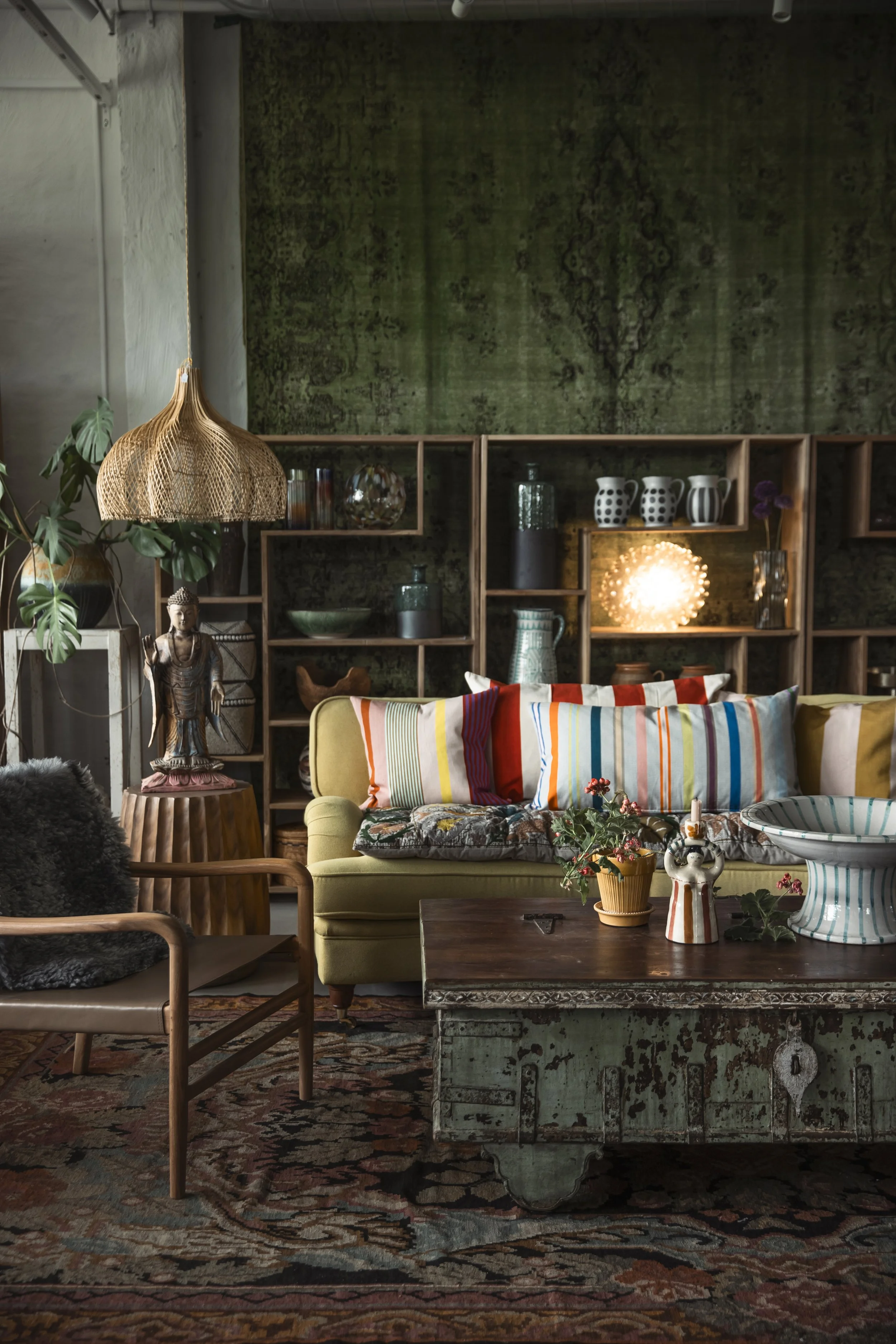 Ett inomhusrum med vardagsrumsmöbler, som en gul soffa med randiga kuddar, en träbord med blomkrukor, en öppen hylla med keramik och glas, en växt, samt en vintage träskåp. Golvet har en stor romsktäckning, och väggarna är decovermålade med grönt och mönster. En ratanlampskärm hänger från taket.