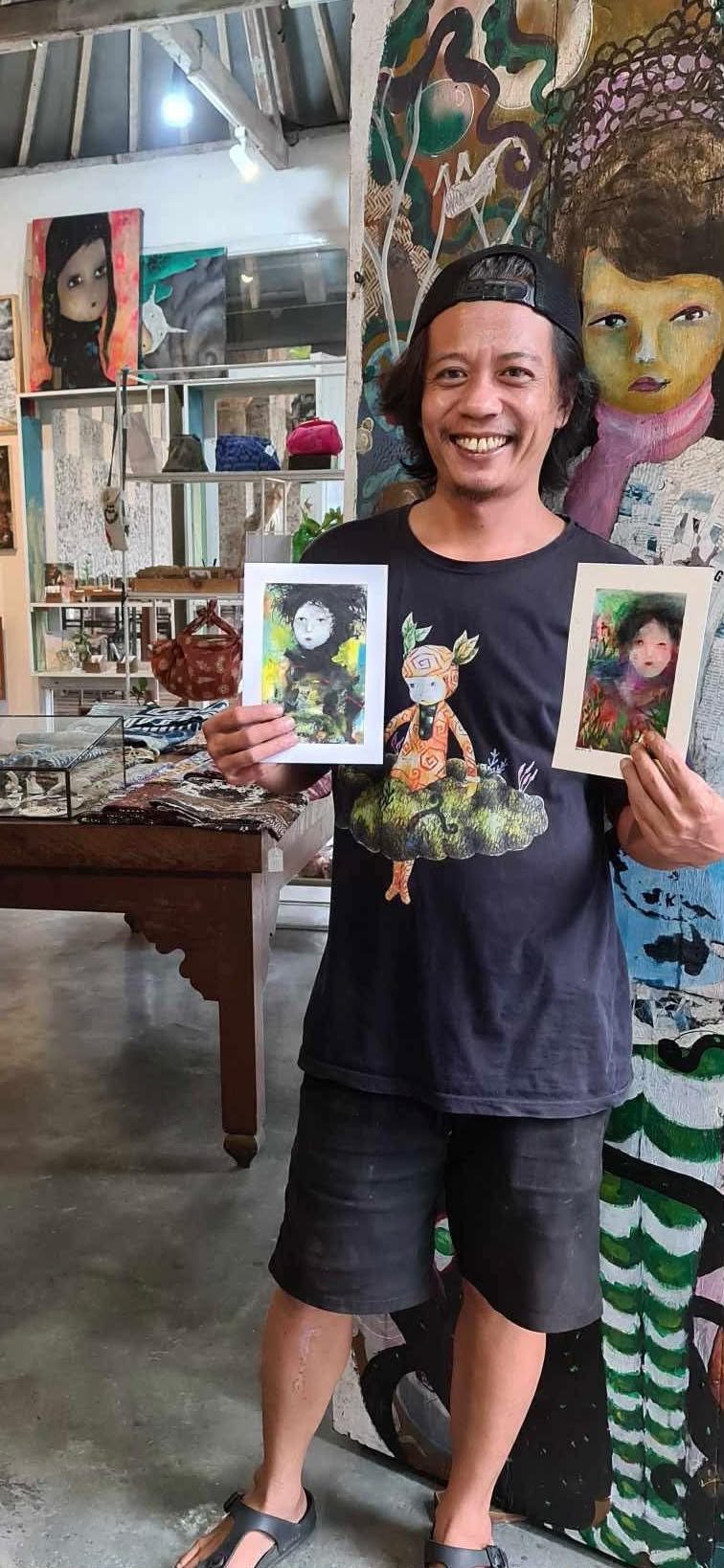 En glad man som visar två små konstverk i händerna, inomhus i ett konstgalleri eller en butik, med stora färgglada målningar på väggarna bakom honom.