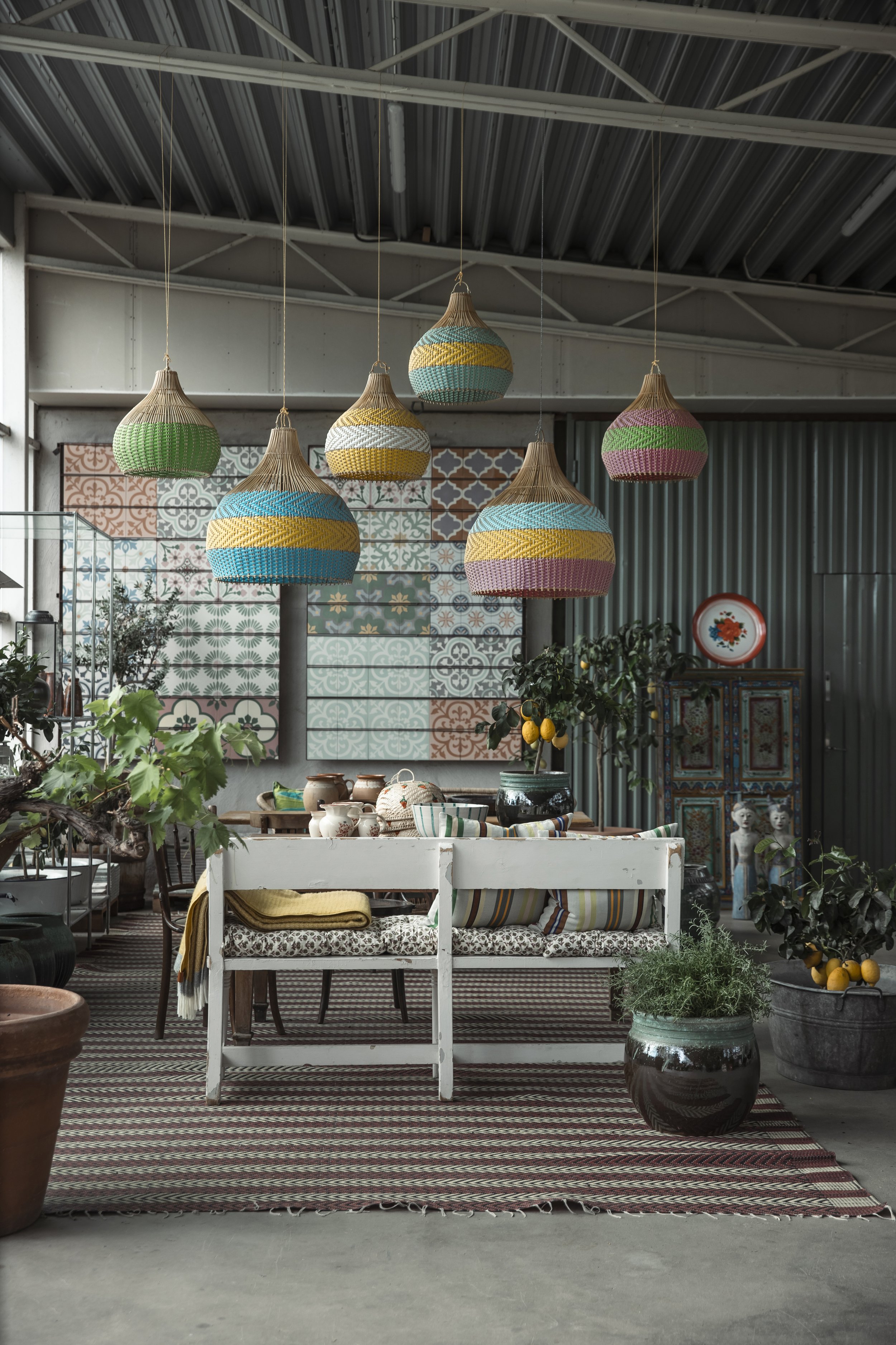 Inomhusmiljö med utsmyckade lampor i taket, ett bord med keramik och textilier, växter i krukor och ett dekorativt skåp i bakgrunden, samt väggar med mönstrade plattor.