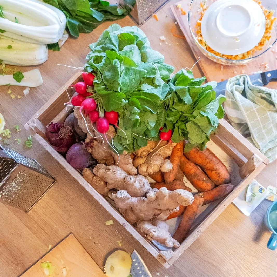 Een houten krat gevuld met diverse groenten zoals radijs, gember, wortels en aardappelen, op een keukentafel.