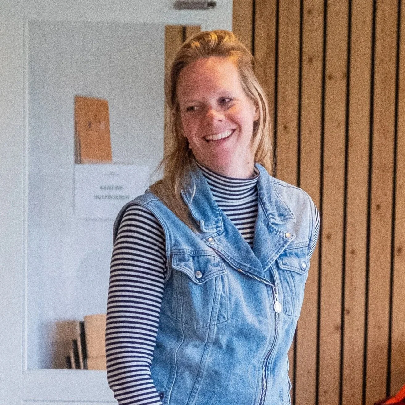 Vrouw met lang blond haar, lachend en gekleed in een spijkerjack en een zwarte-wit gestreepte trui, binnen