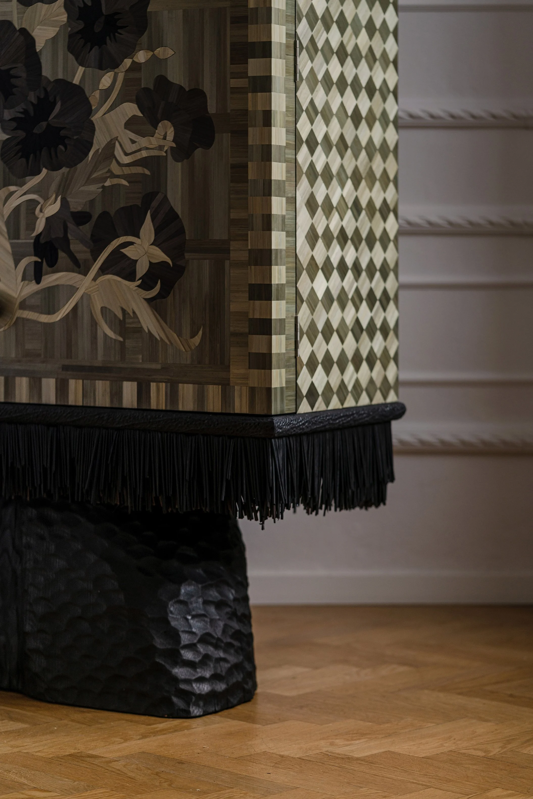 Close-up of an intricately designed wooden cabinet with floral inlay and a black textured base, on a wooden floor.