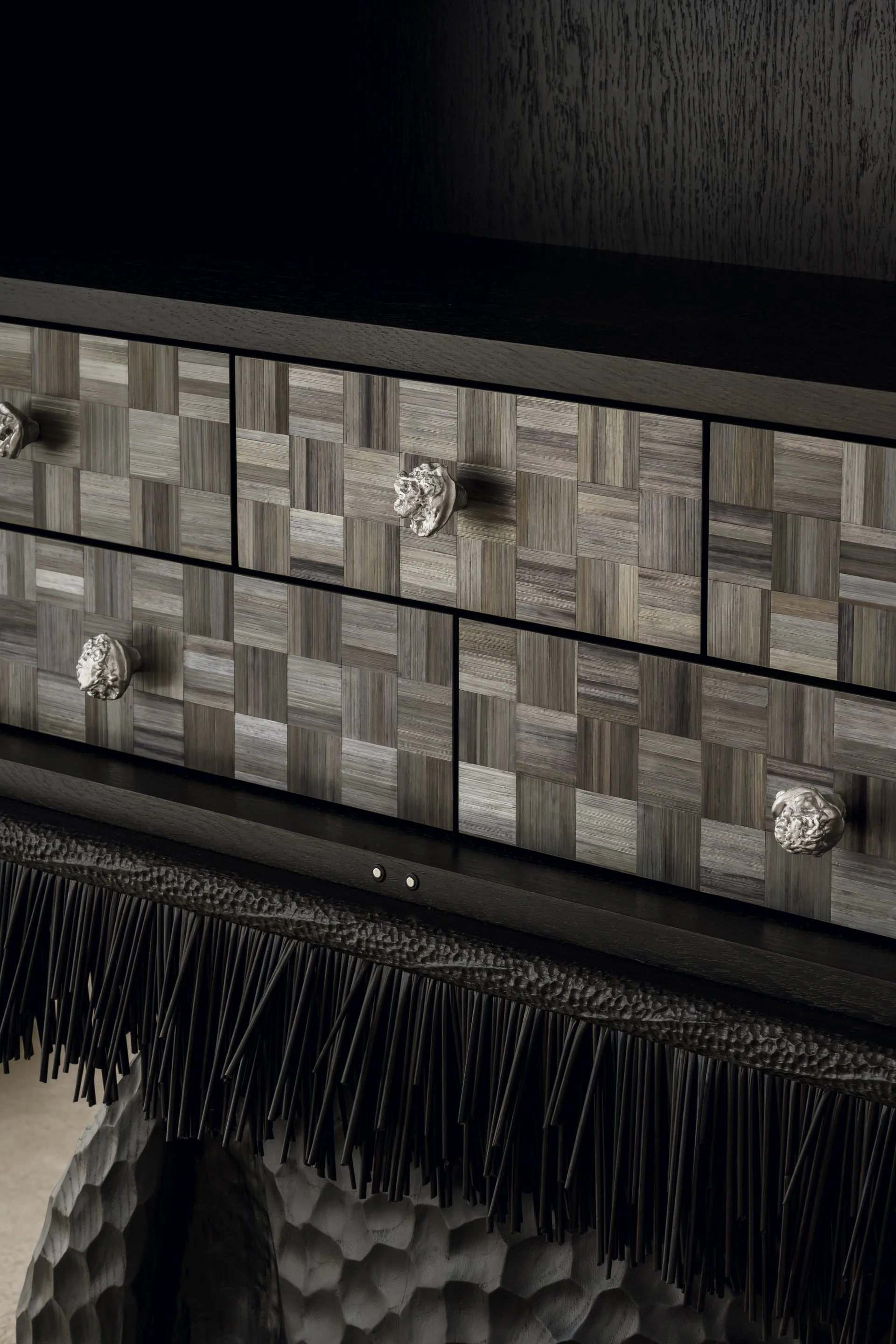Close-up of a modern black wooden dresser with a woven geometric pattern on the drawers and ornate silver handles, with a textured black fringe on the lower edge.