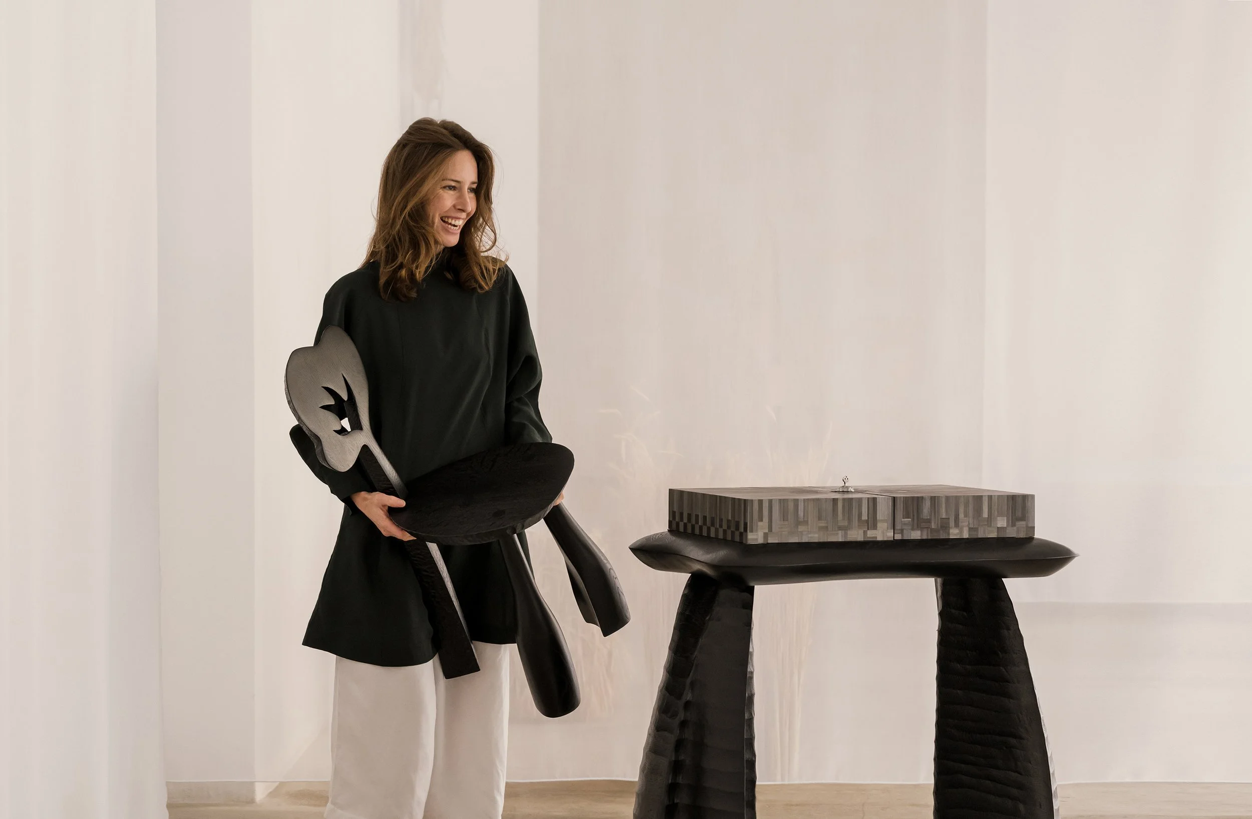 A woman with brown hair, wearing a black top and white pants, smiling while holding a black sculpture of a face and a rounded base, standing next to a wooden table with a geometric patterned object on top in a minimalist white room.
