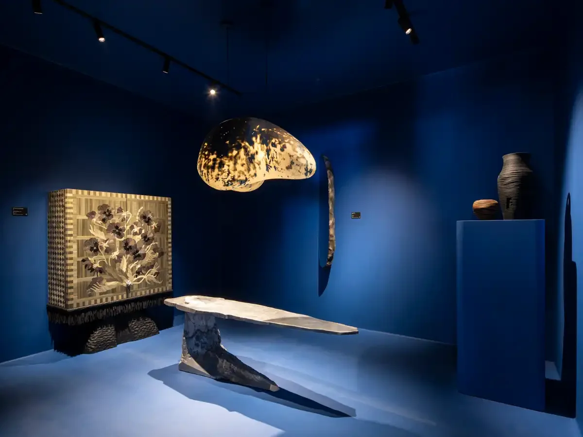 An art gallery with blue walls displaying various sculptures and art pieces, including a large abstract sculpture hanging from the ceiling, a tree-themed artwork on the wall, a prominent marble or stone bench, and a pair of vases on a pedestal.