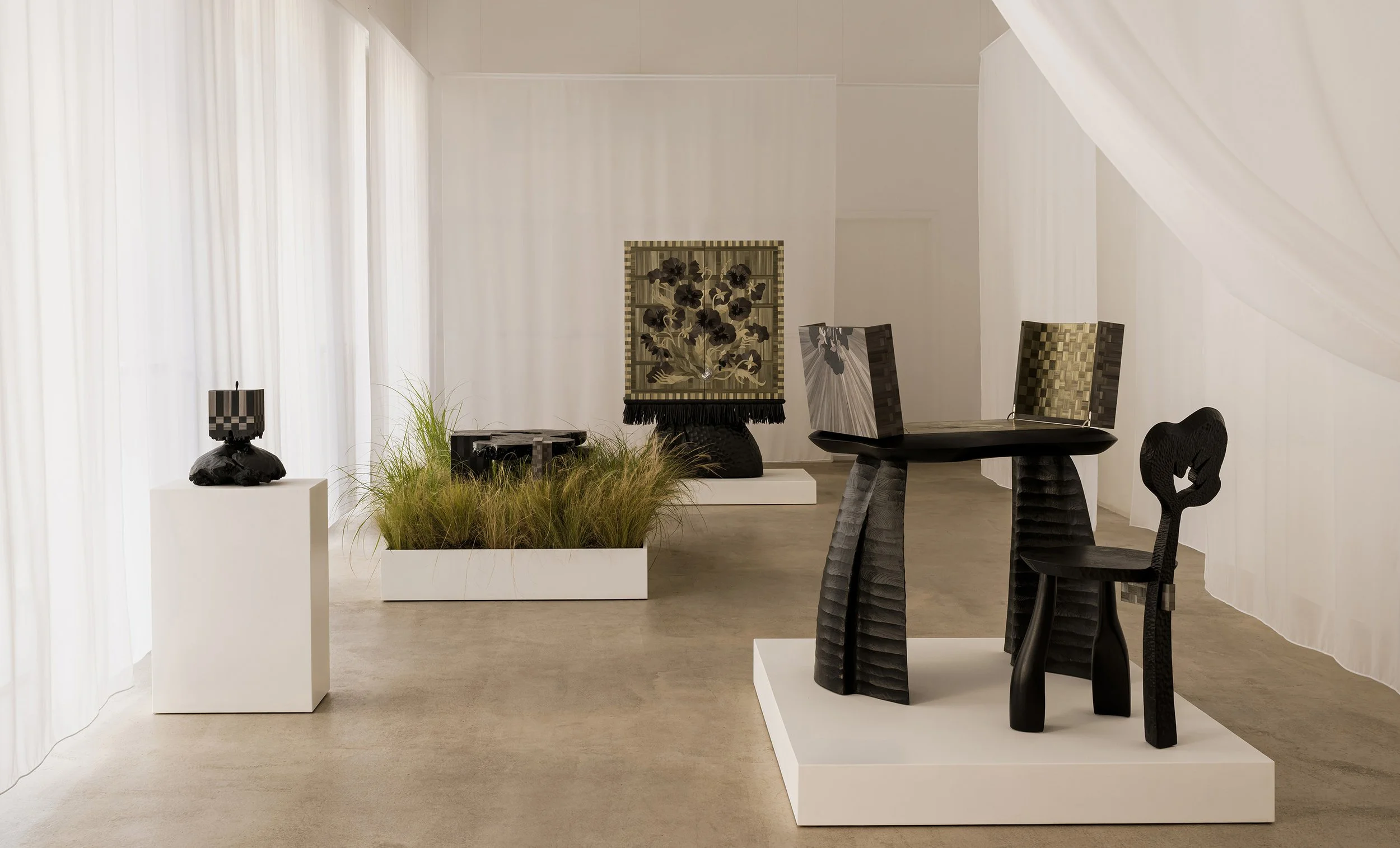 An art gallery with various black sculptural furniture and artwork displayed on pedestals, with sheer white curtains and soft lighting.