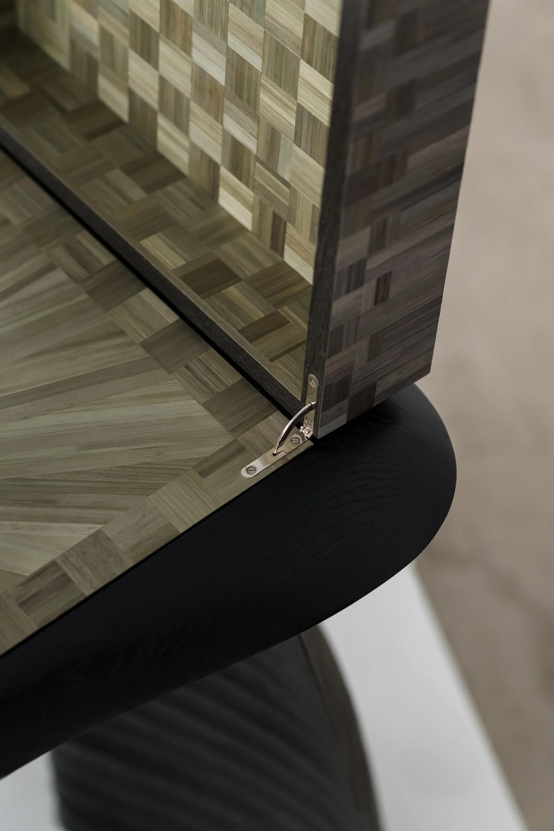 Close-up of a corner of a table with a mosaic-style top, featuring small square wooden tiles in various shades of beige and gray, with metal hinges attaching a decorative wooden panel to the edge.
