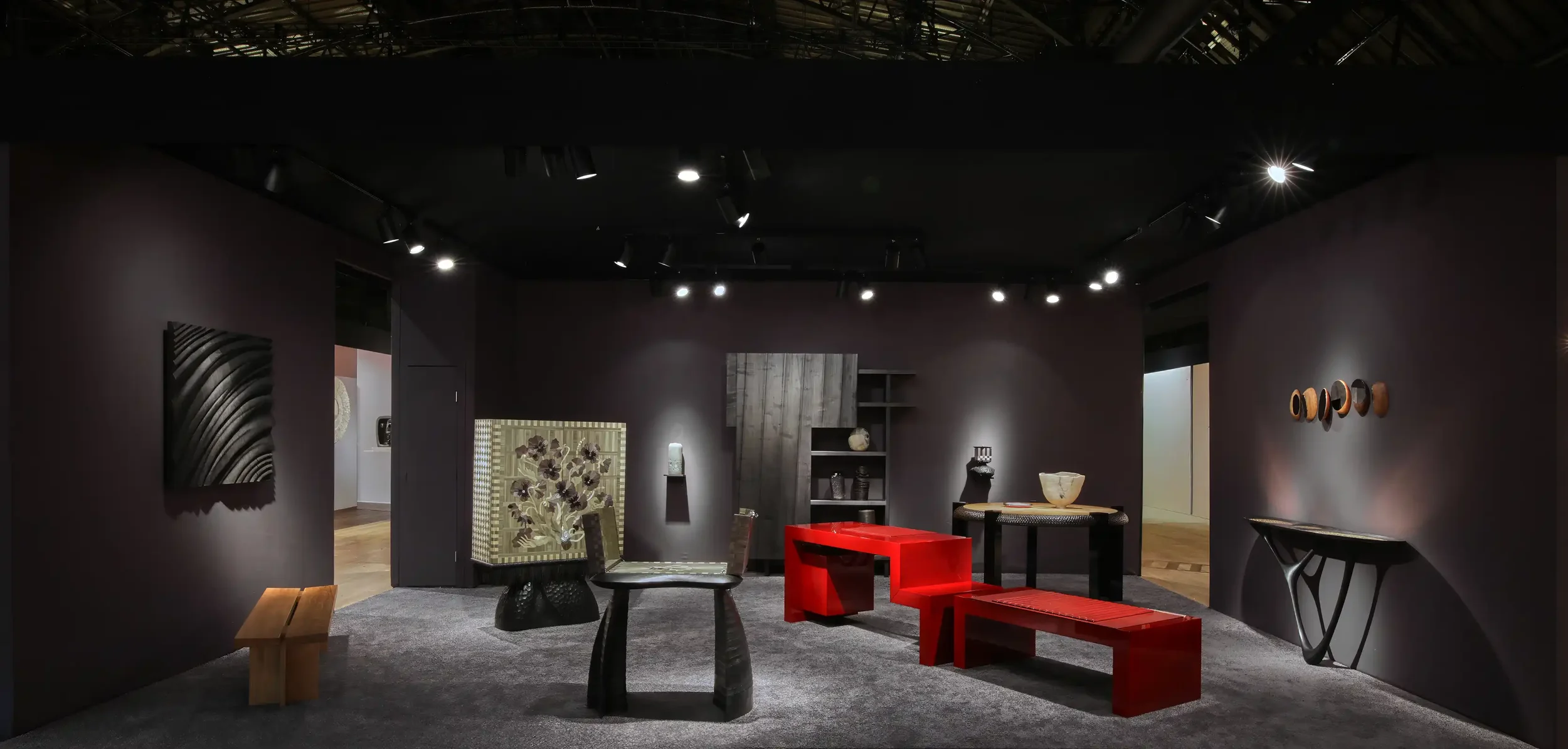 An art gallery with dark walls and track lighting, displaying modern furniture and decorative objects, including a red bench, black chairs, a side table, and wall-mounted sculptures.