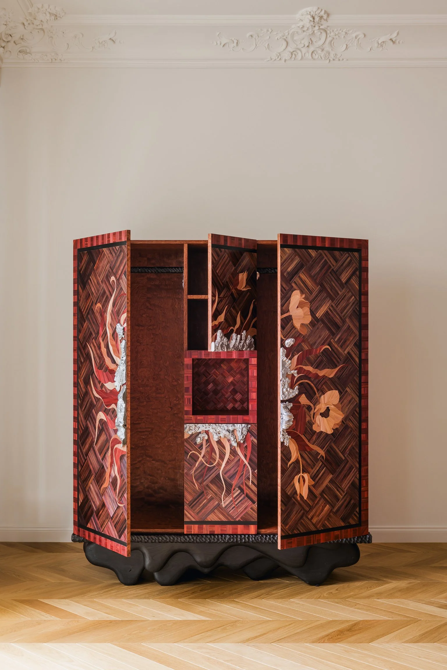 A wooden cabinet with intricate floral and geometric inlay designs, including flowers, leaves, and a central square pattern, set against a plain wall with decorative molding.