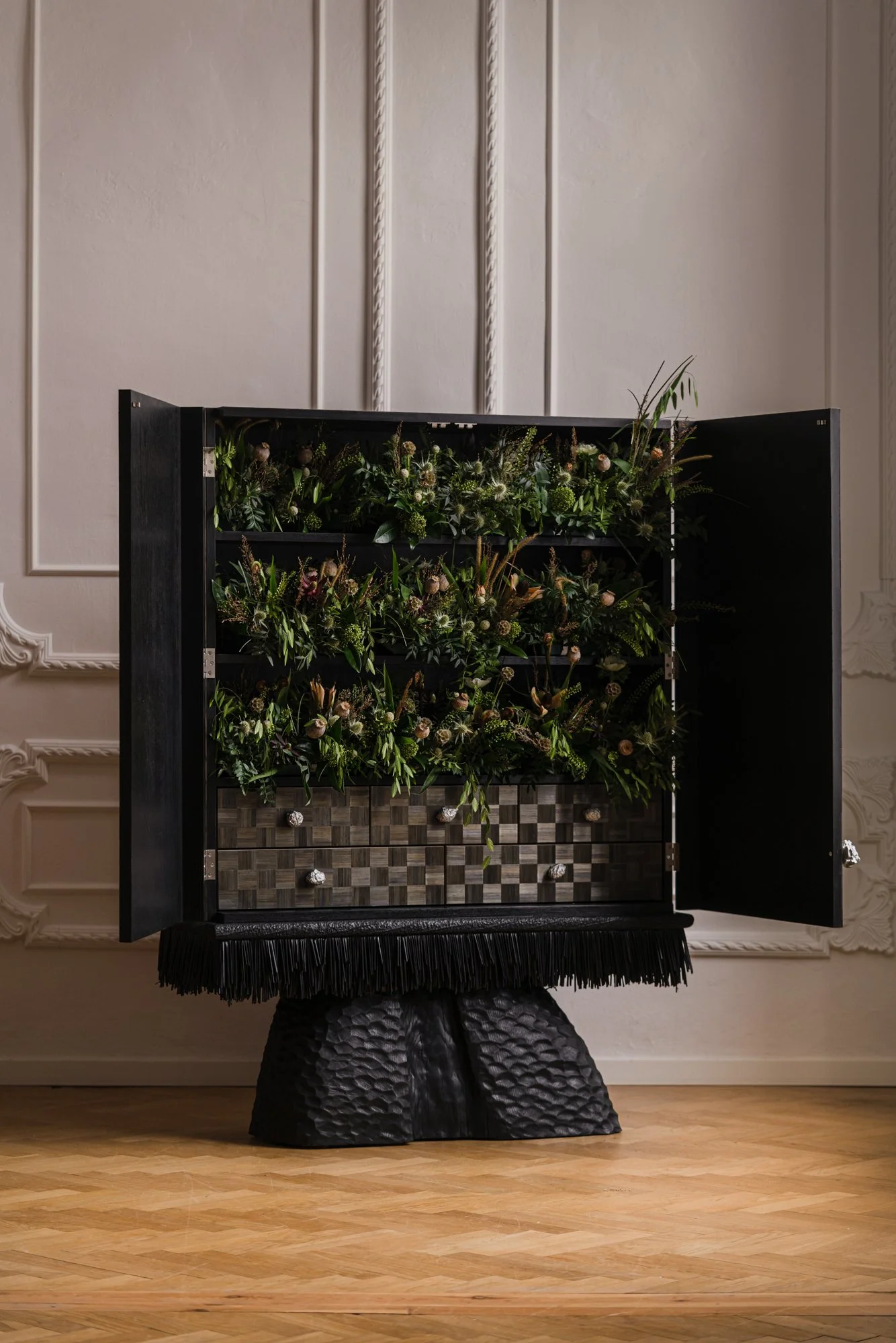 A black display cabinet with floral arrangements inside, standing on a textured black base, positioned against a beige wall with decorative moldings.