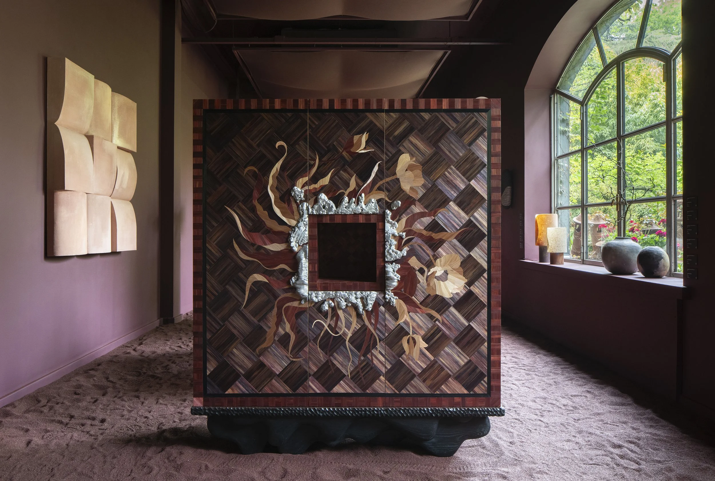 Decorative cabinet with geometric pattern and floral design, situated in a room with large arched window, pink walls, and artwork on the wall.