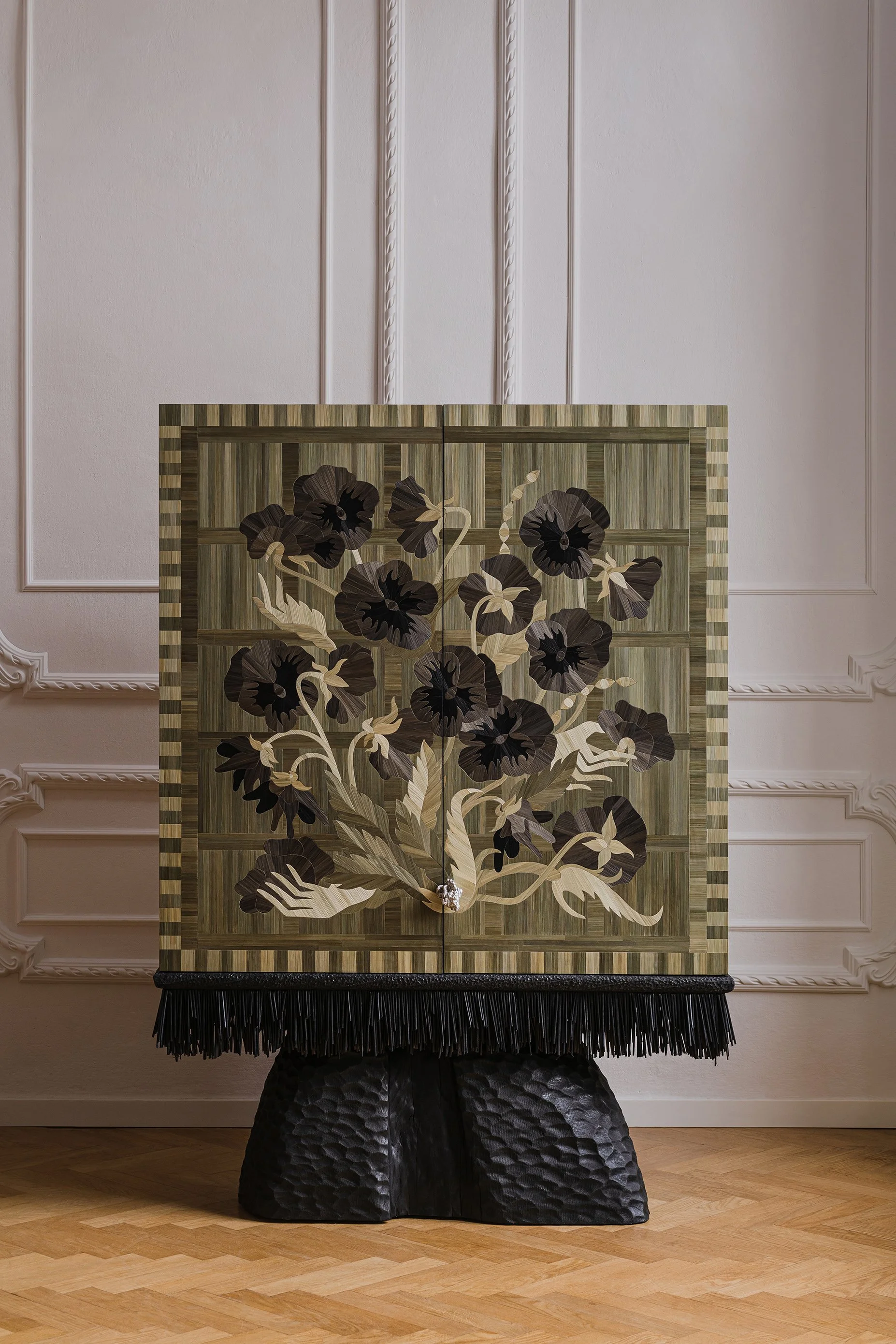 A decorative wooden cabinet with floral inlay design, featuring dark and light-colored flowers and leaves, topped with a fringe, and supported by a textured black base, set against a white paneled wall with intricate molding details.