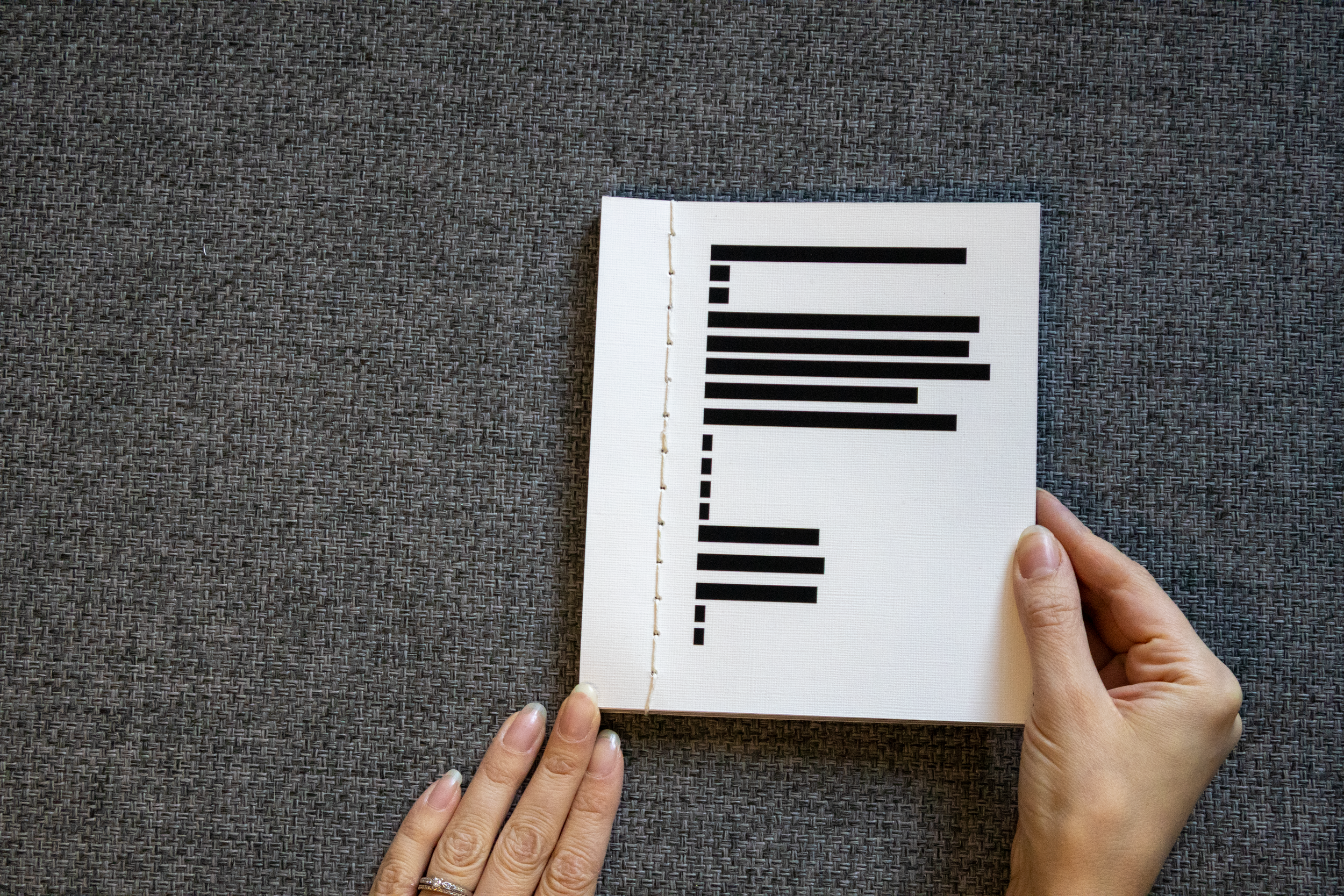 Livre ouvert avec des lignes de texte en noir, placé sur une surface en tissu gris, avec une main tenant la page droite et une autre la page gauche.