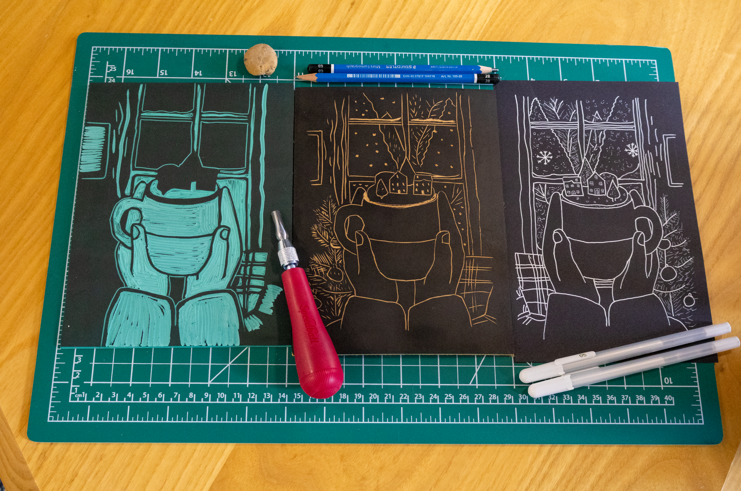 Trois dessins de tasses de Noël, représentés sur deux feuilles noires, avec des détails de décorations hivernales, un crayon, deux marqueurs, un pinceau, un objet rond, et deux stylos blancs, sur une planche de découpe verte sur un bureau en bois.