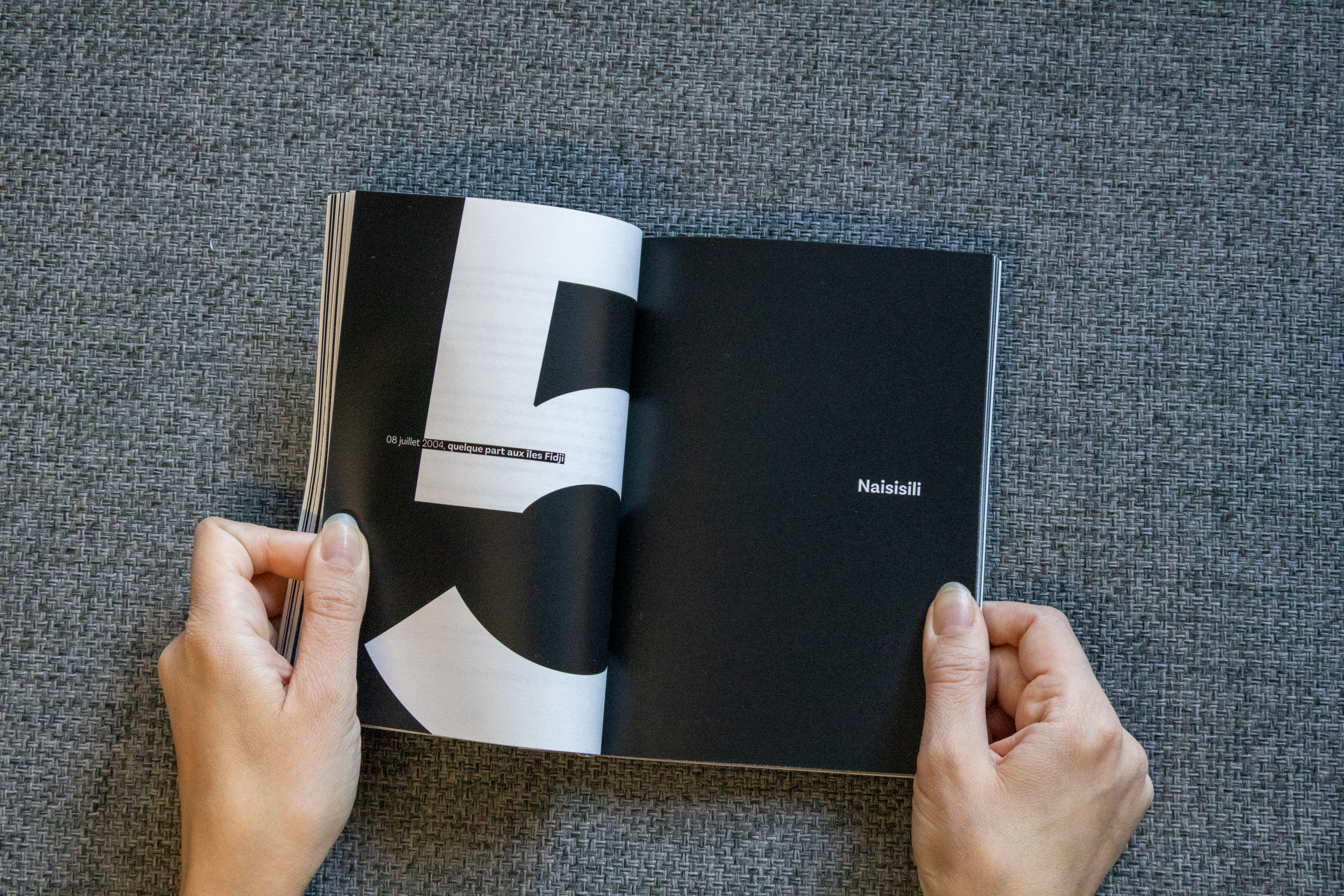 Une personne tient un livre ouvert sur un fond de tissu gris, avec une page noire avec du texte en français et une autre page noire avec le mot 'Naisilili' écrit en blanc.