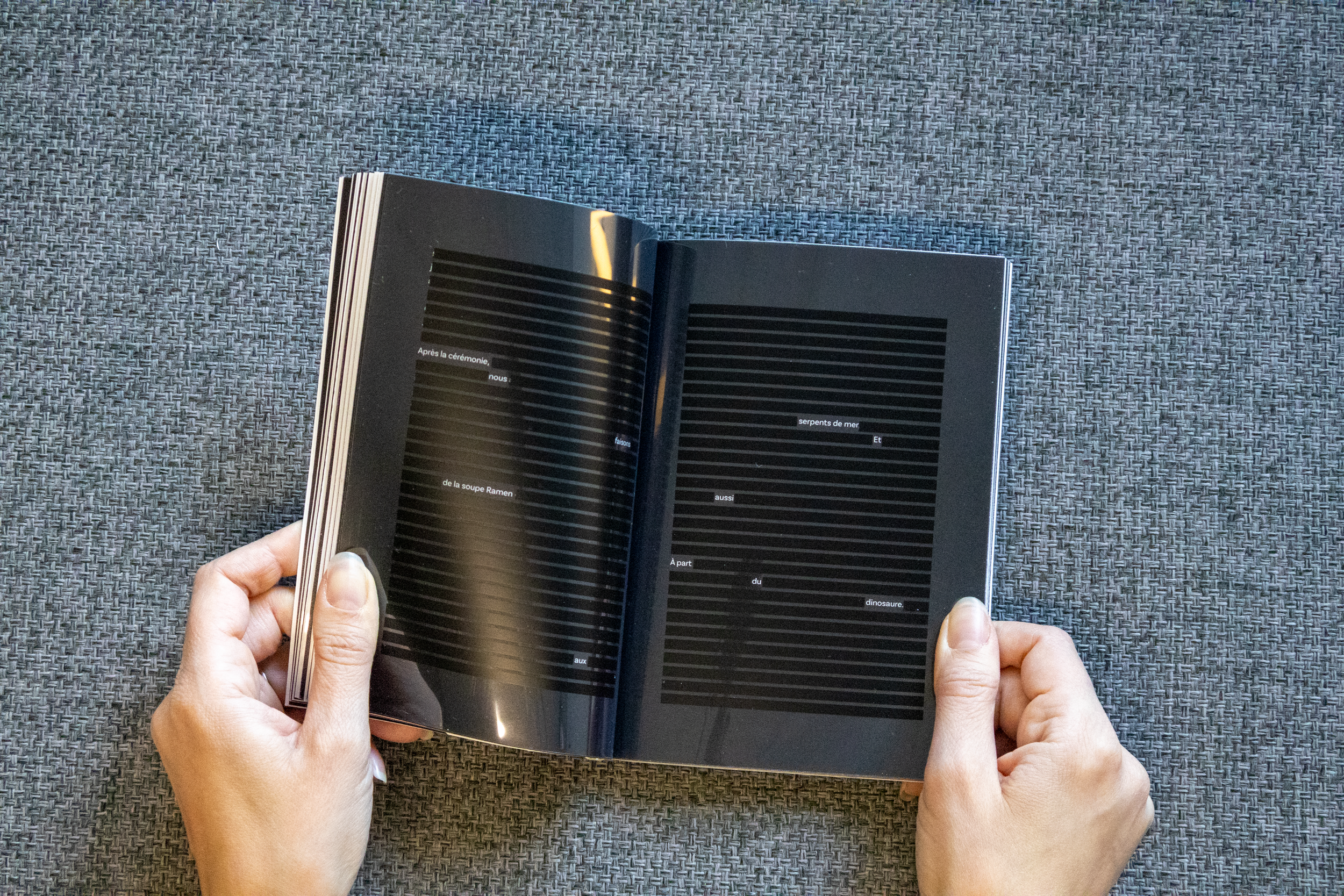 Une personne tient un livre ouvert à deux mains sur une surface en tissu gris, la page montre un texte en français sur un fond noir avec des lignes horizontales.