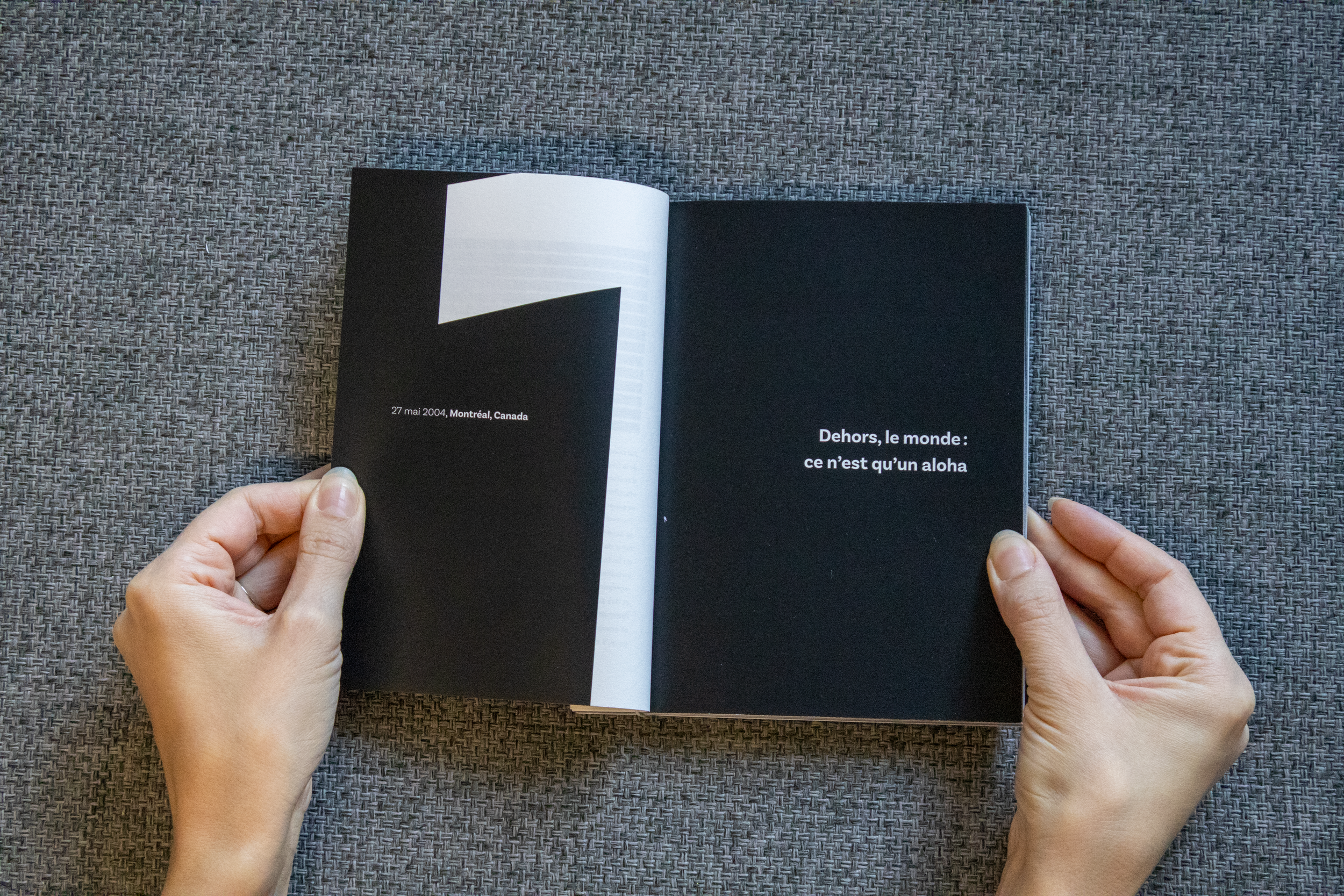 Une personne tient un livre ouvert avec deux pages noires. La page de gauche affiche la date '27 mai 2004' et l'emplacement 'Montréal, Canada'. La page de droite contient le texte 'Dehors, le monde : ce n’est qu’un aloha'. Le livre est placé sur une 