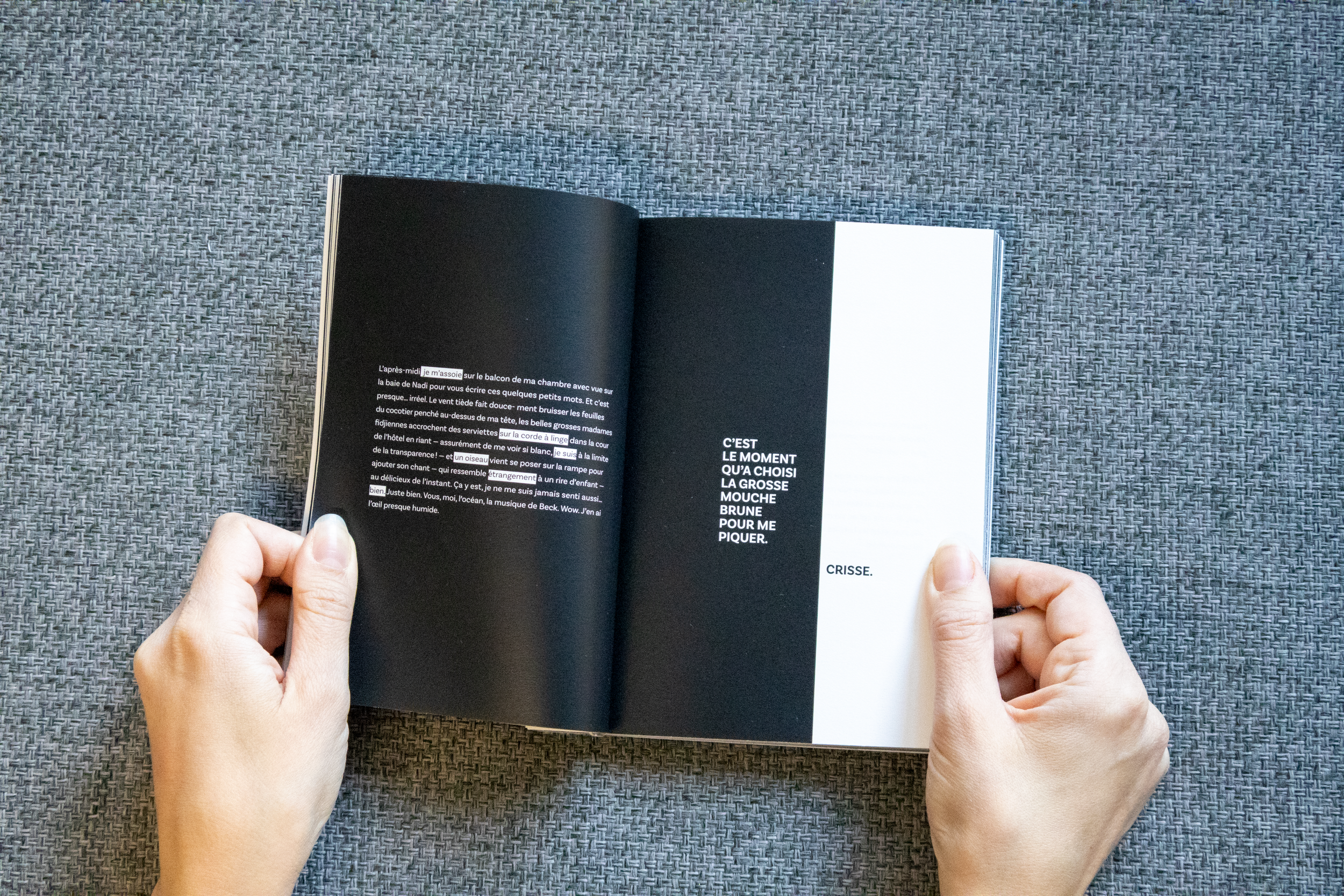 Une personne tenant un livre ouvert avec une mise en page artistique, avec du texte en français sur fond noir et blanc, posé sur une surface en tissu gray.