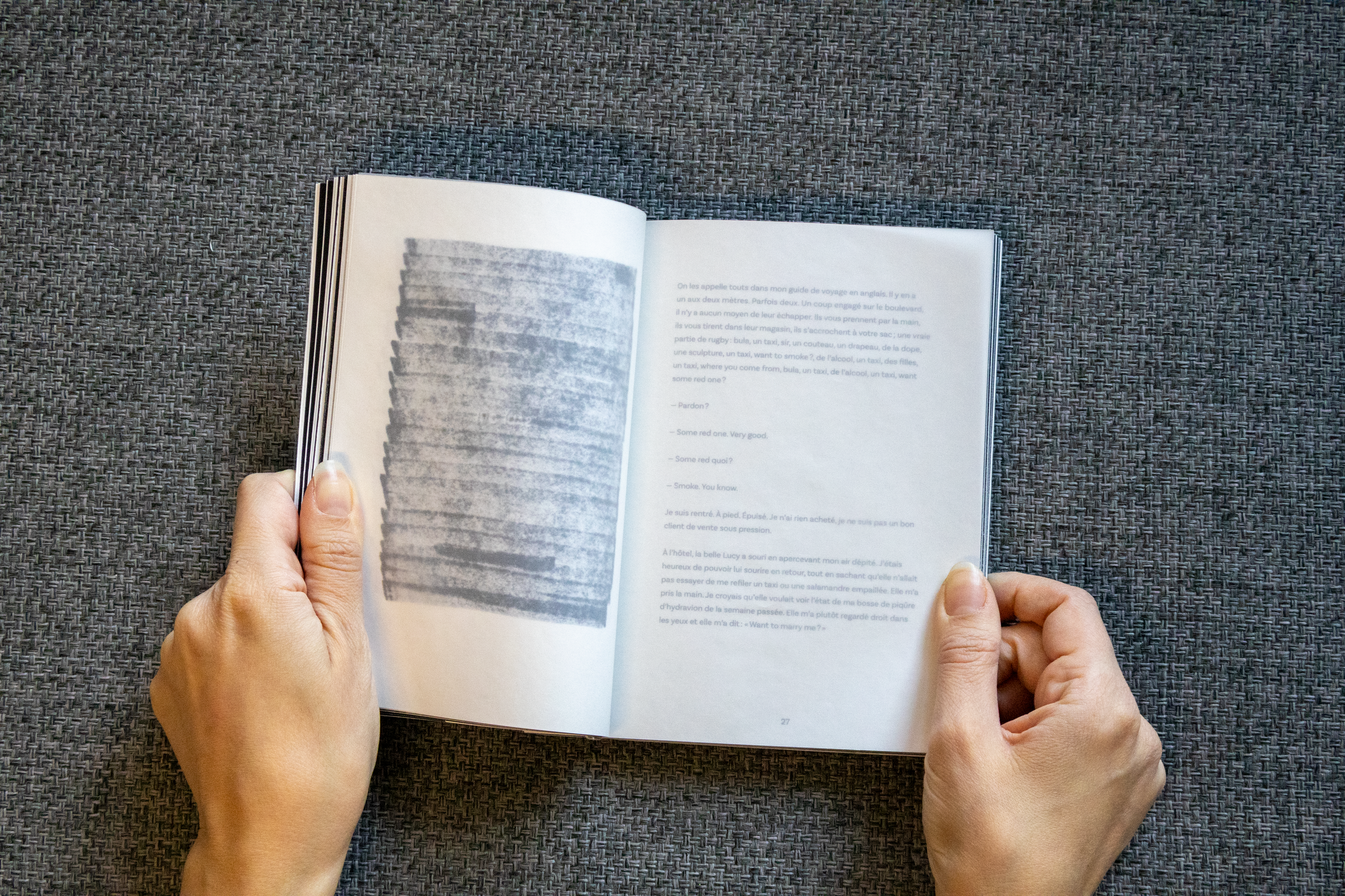 Une personne tient un livre ouvert avec des pages en français, posé sur une surface en tissu gris.