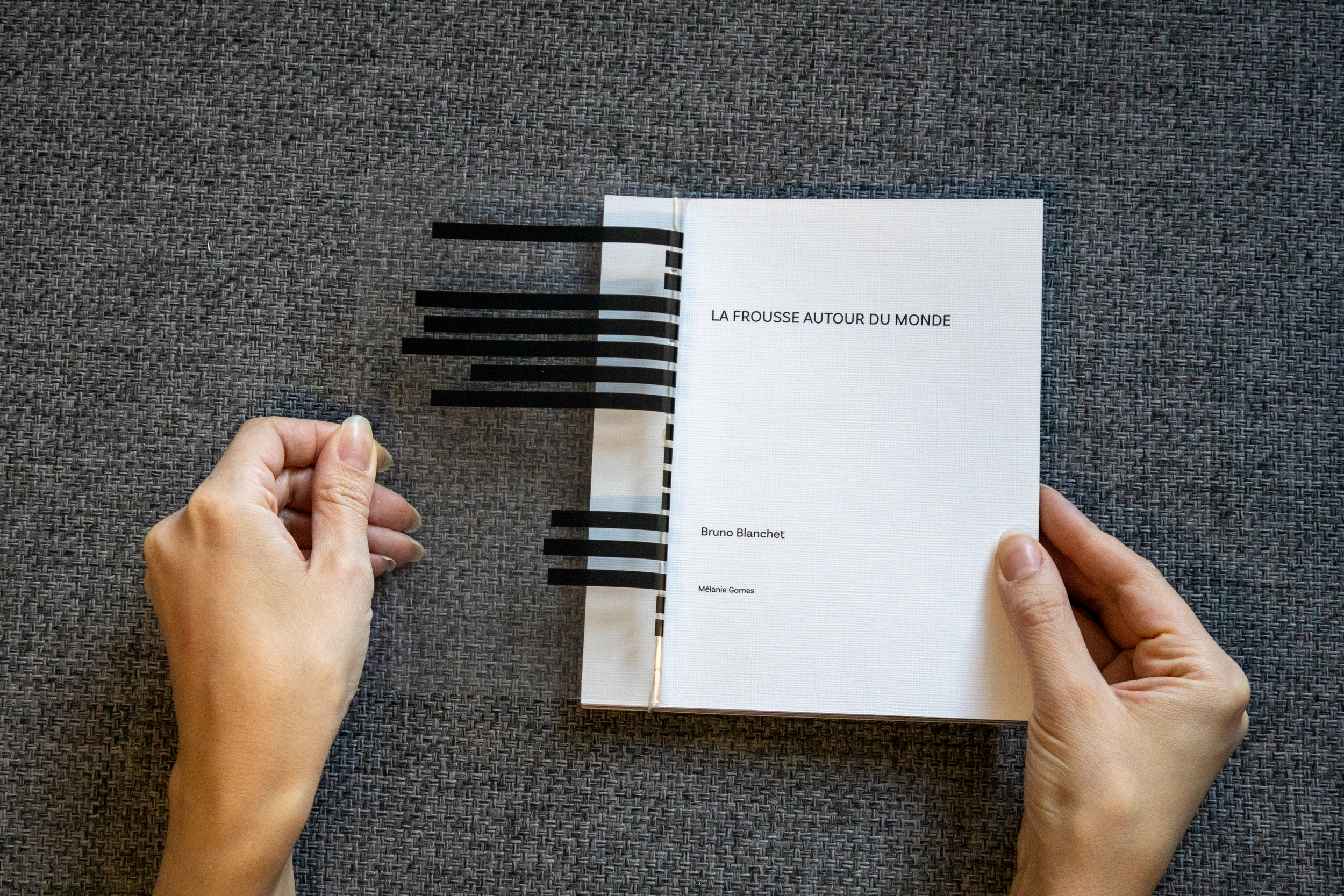 Page de couverture d'un livre intitulé 'La Frouss autour du Monde' avec le nom d'auteurs Bruno Blanchet et Mélanie Gomes, posé sur une surface en tissu gris, avec un marque-page noir à rayures horizontales.