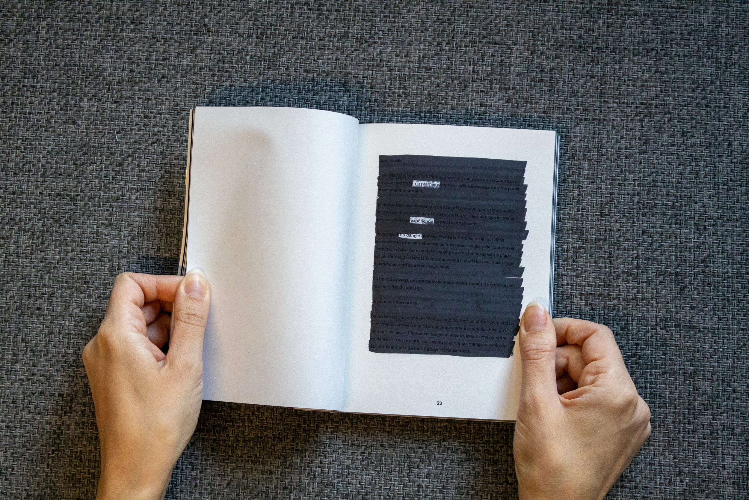 Une personne tient un livre ouvert sur une surface en tissu gris, avec du texte partiellement masqué par correction.