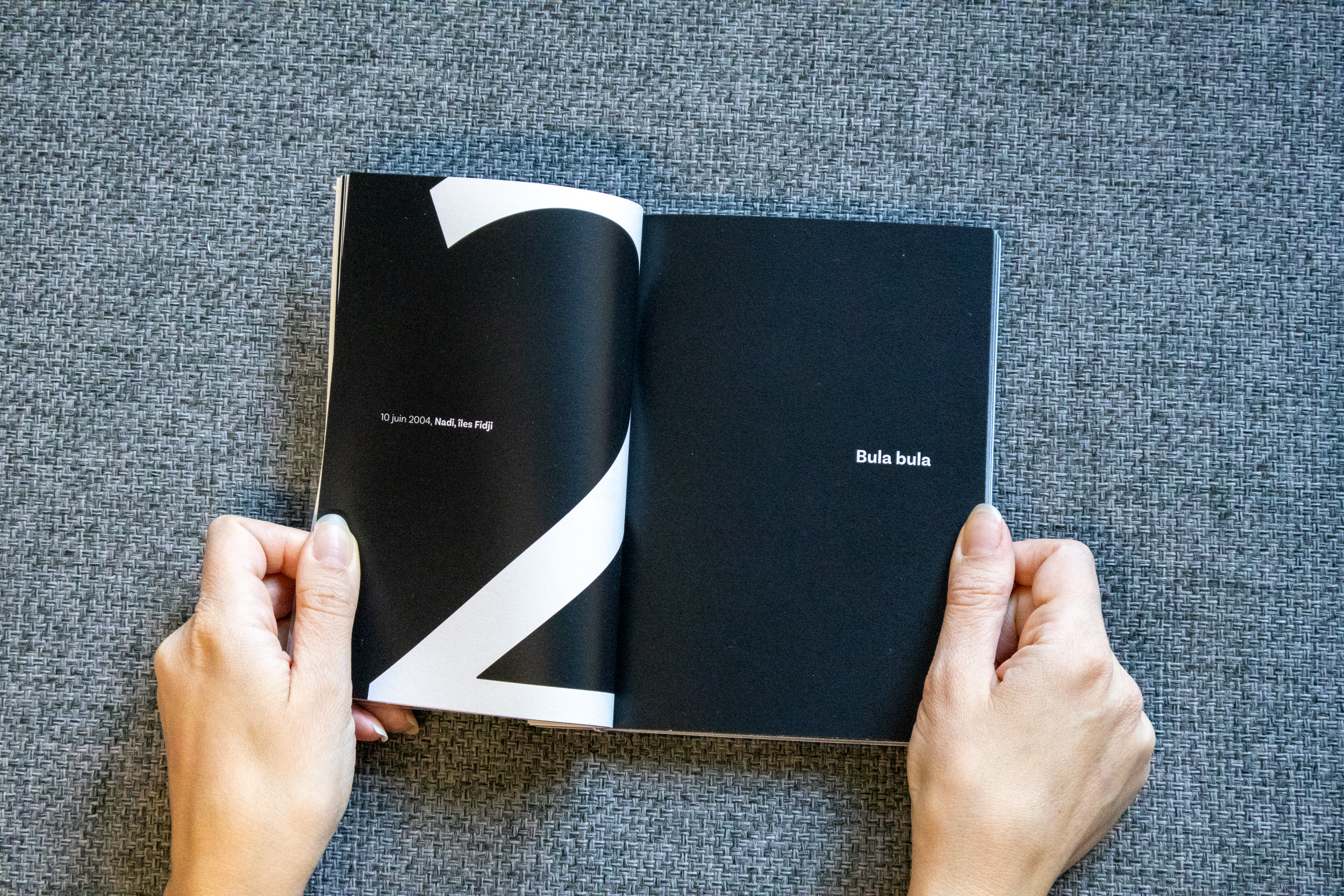 Livre ouvert avec une double page, la page gauche montre un texte en français "10 juin 2004, Nadi, îles Fidji" sur un fond noir et blanc abstrait, la page droite affiche le texte "Bula bula" sur un fond noir.