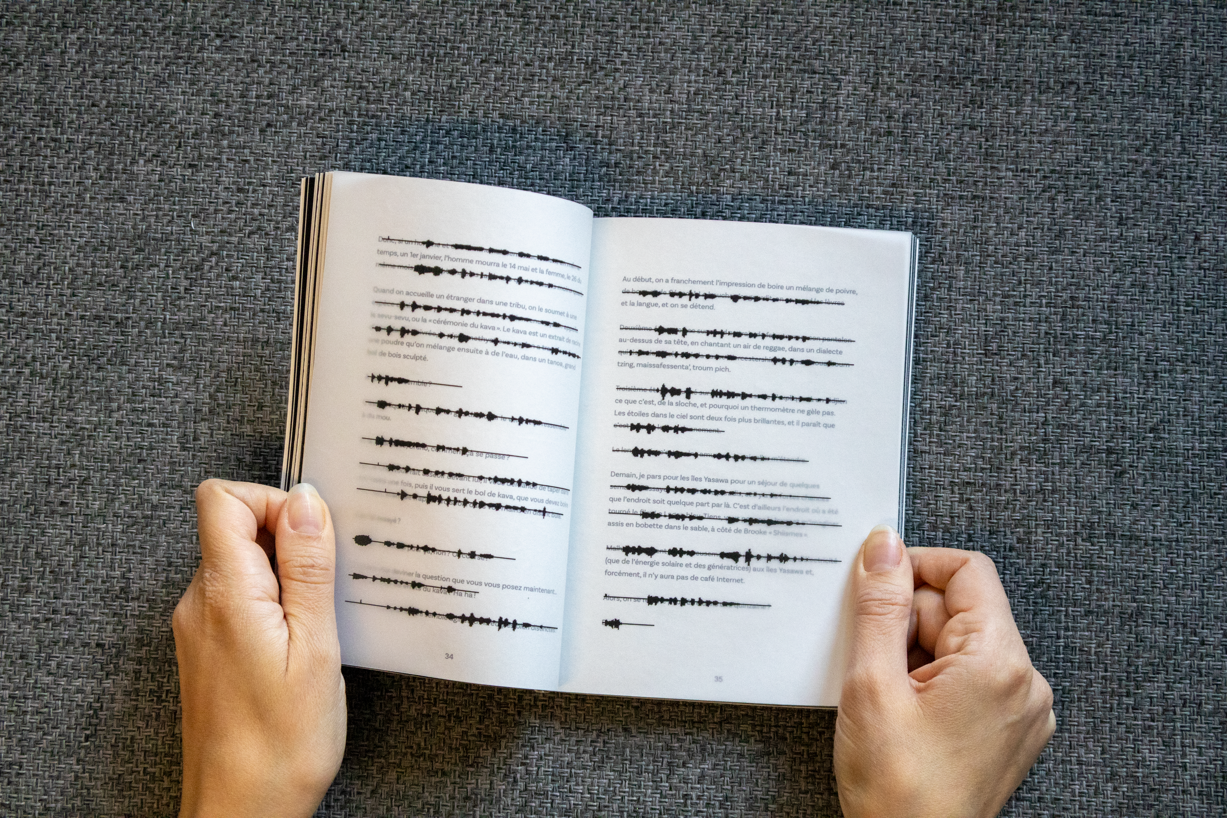 Un livre ouvert avec des pages contenant des marques noires de ciseaux à la place du texte, tenu par deux mains, posé sur une surface en tissu gris.